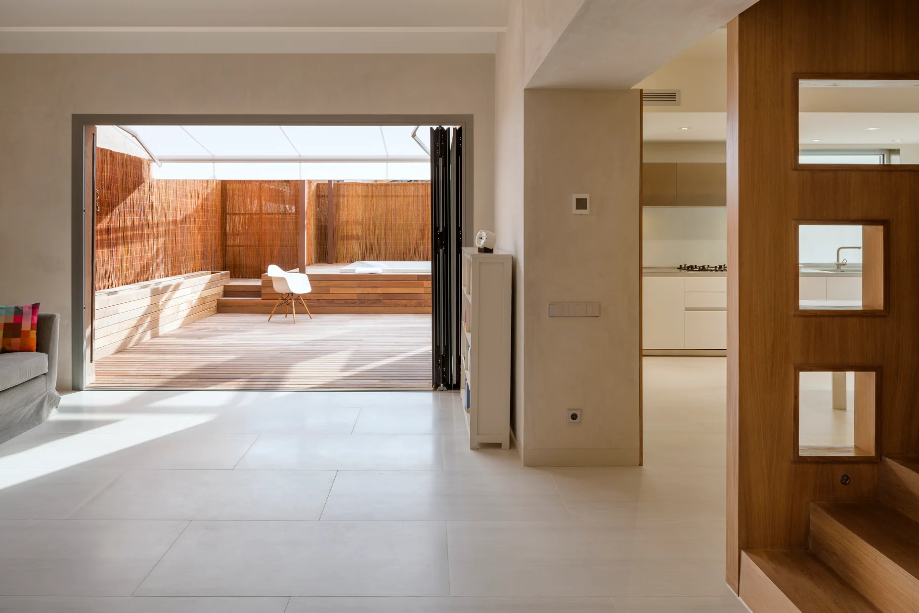 A bright minimalist living room features large floor tiles, a folding glass door leading to a wooden patio deck, and a wood partition wall.
