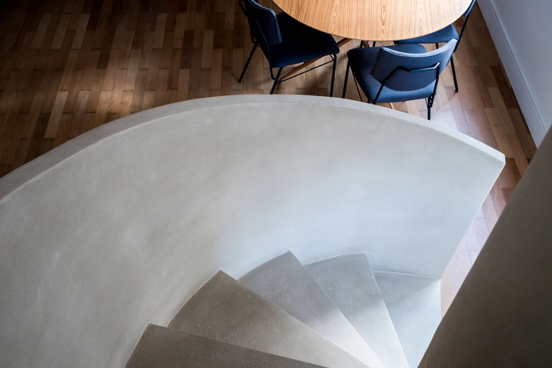 A high-angle view of a sculpted, curved concrete staircase next to a round wooden dining table with dark blue upholstered chairs.