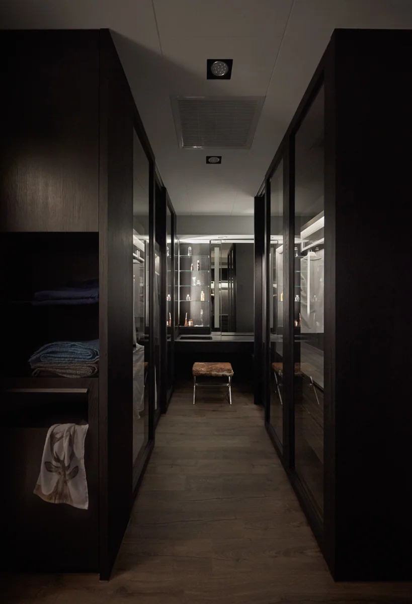 A dark-toned walk-in closet featuring floor-to-ceiling glass-front wardrobes, recessed ceiling lights, and a small upholstered stool at the far end.