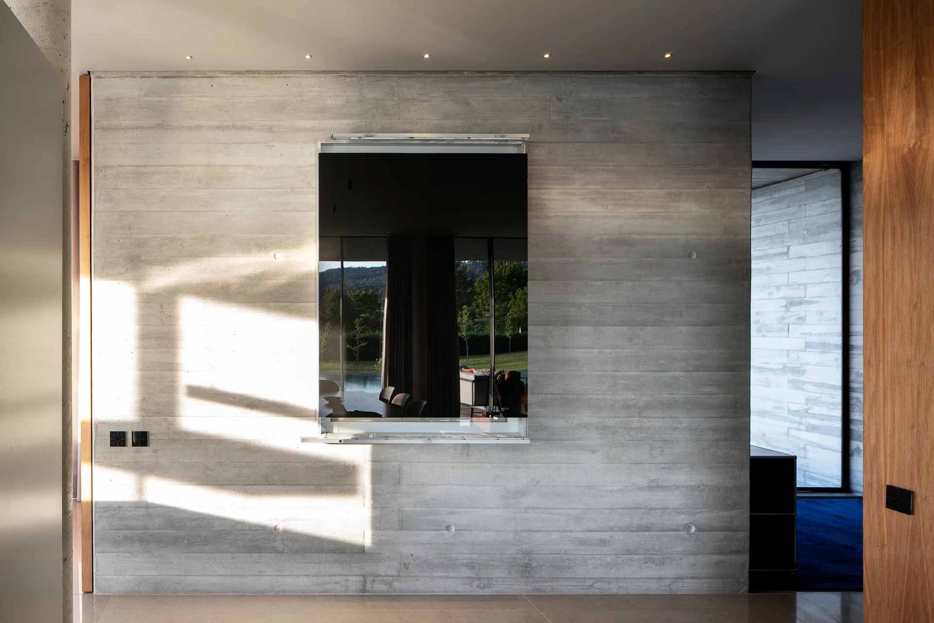 A rectangular wall-mounted mirror hangs on a board-formed concrete wall illuminated by sunlight and recessed ceiling spotlights.