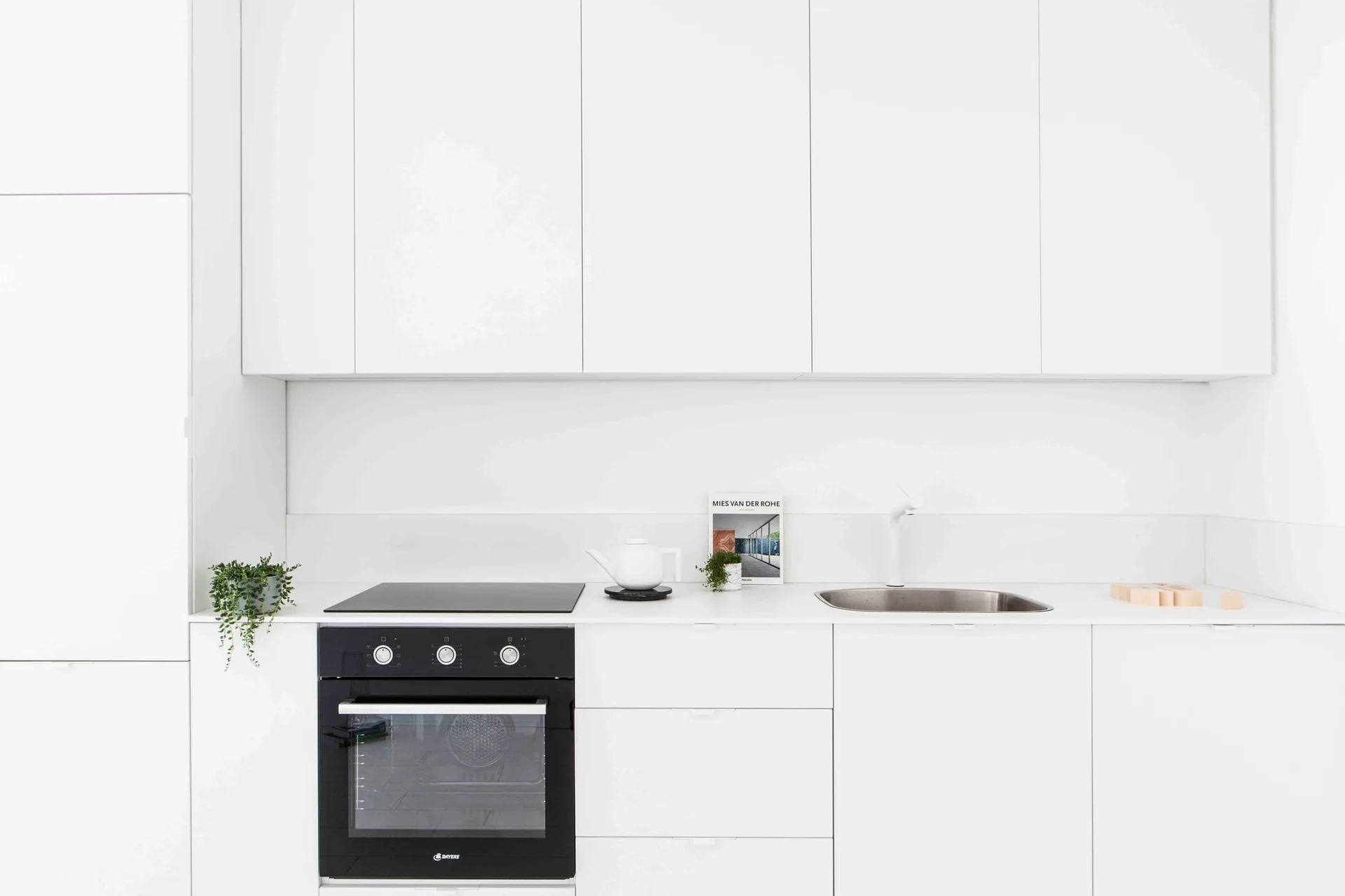 A minimalist white kitchen features handleless upper and lower cabinets, a black built-in oven, and a small stainless steel sink with a white faucet.