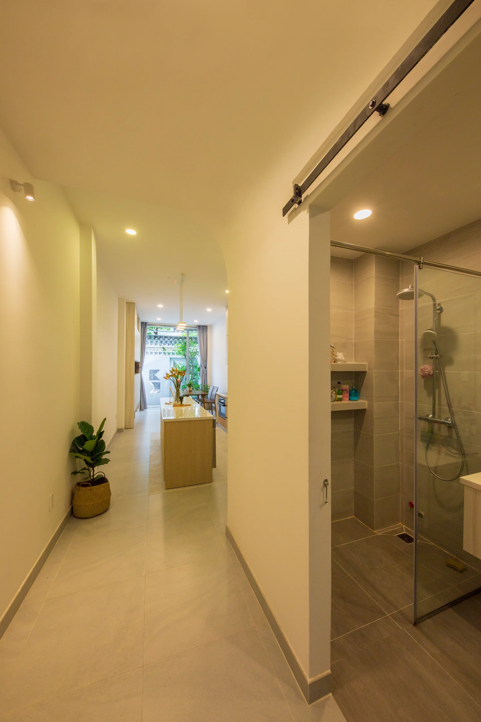 A hallway with grey tiled floors features a glass shower enclosure on the right and leads through an archway to a kitchen island.