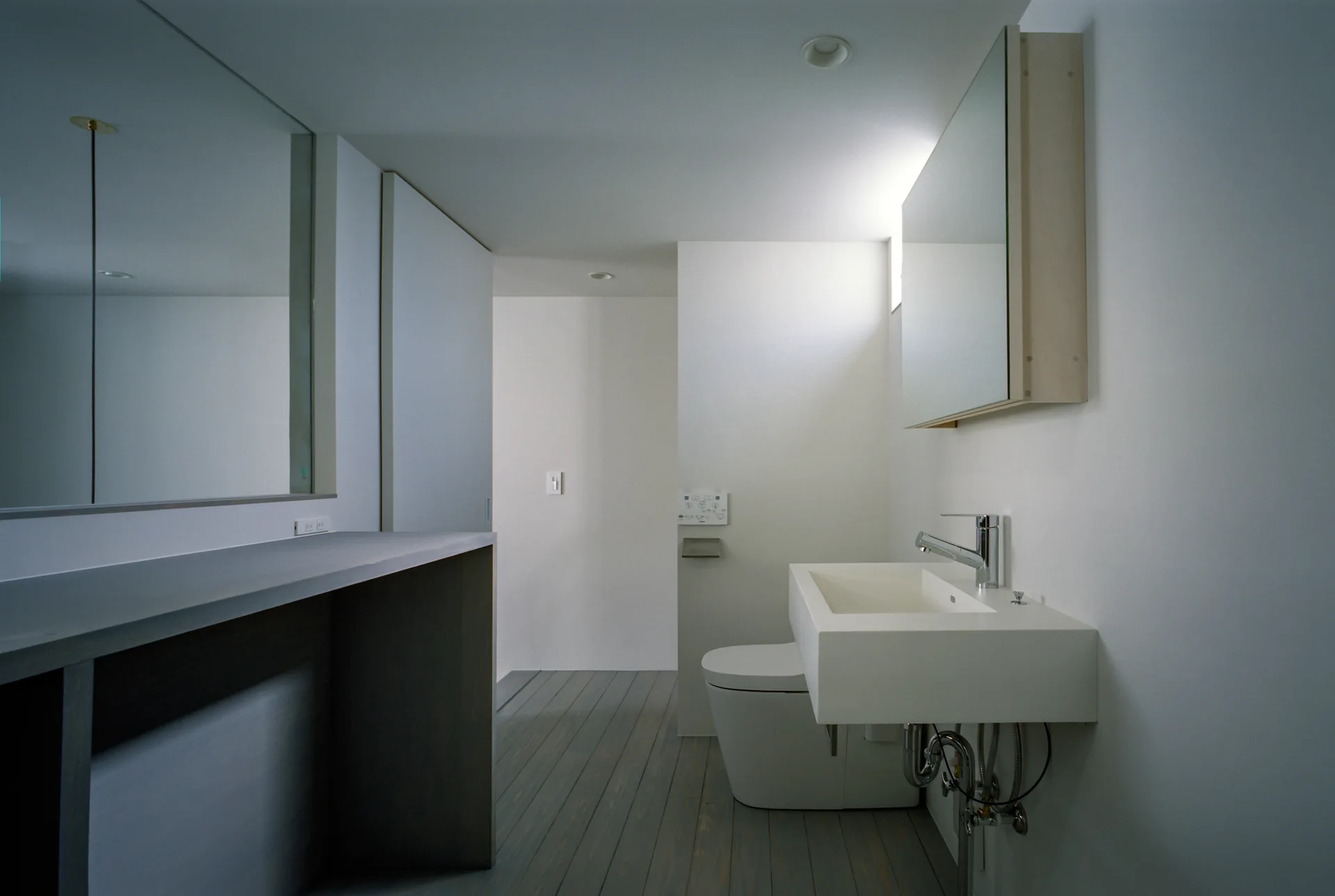 A minimalist bathroom features a wall-mounted rectangular white sink, a modern toilet, a large mirror, and a grey wood plank floor.