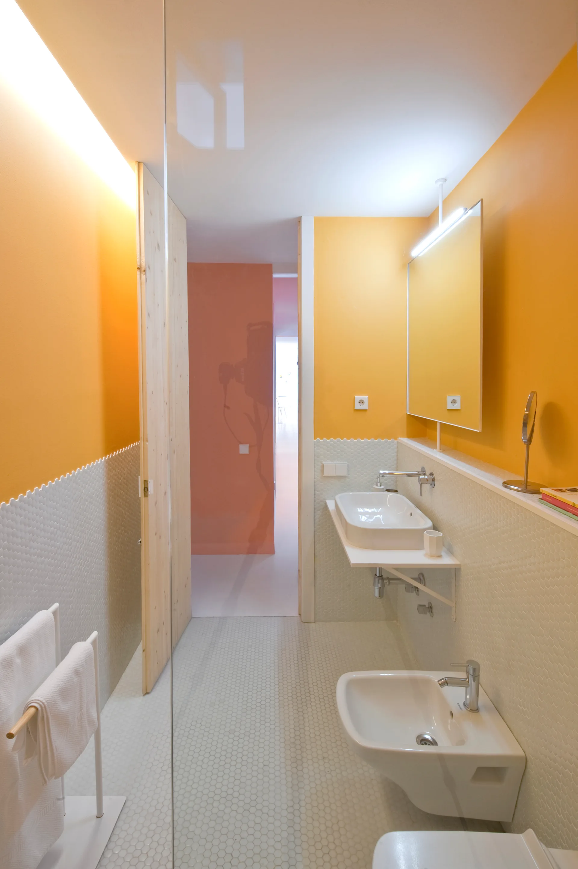 A narrow bathroom features vibrant yellow upper walls, small white hexagonal tiled lower walls and flooring, a floating white sink, and a bidet.