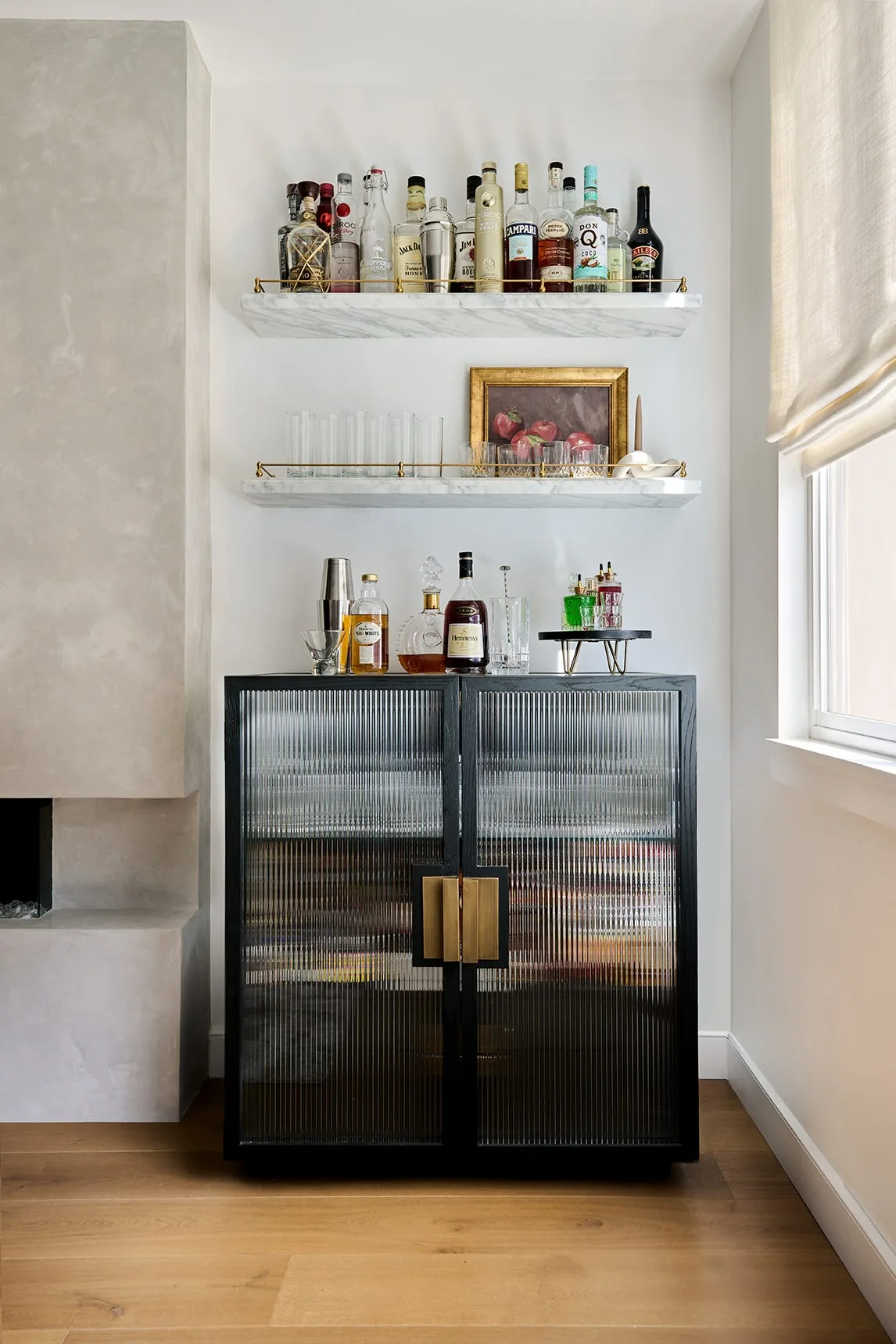 A modern home bar setup featuring a black cabinet with fluted glass doors underneath floating marble shelves stocked with liquor bottles.