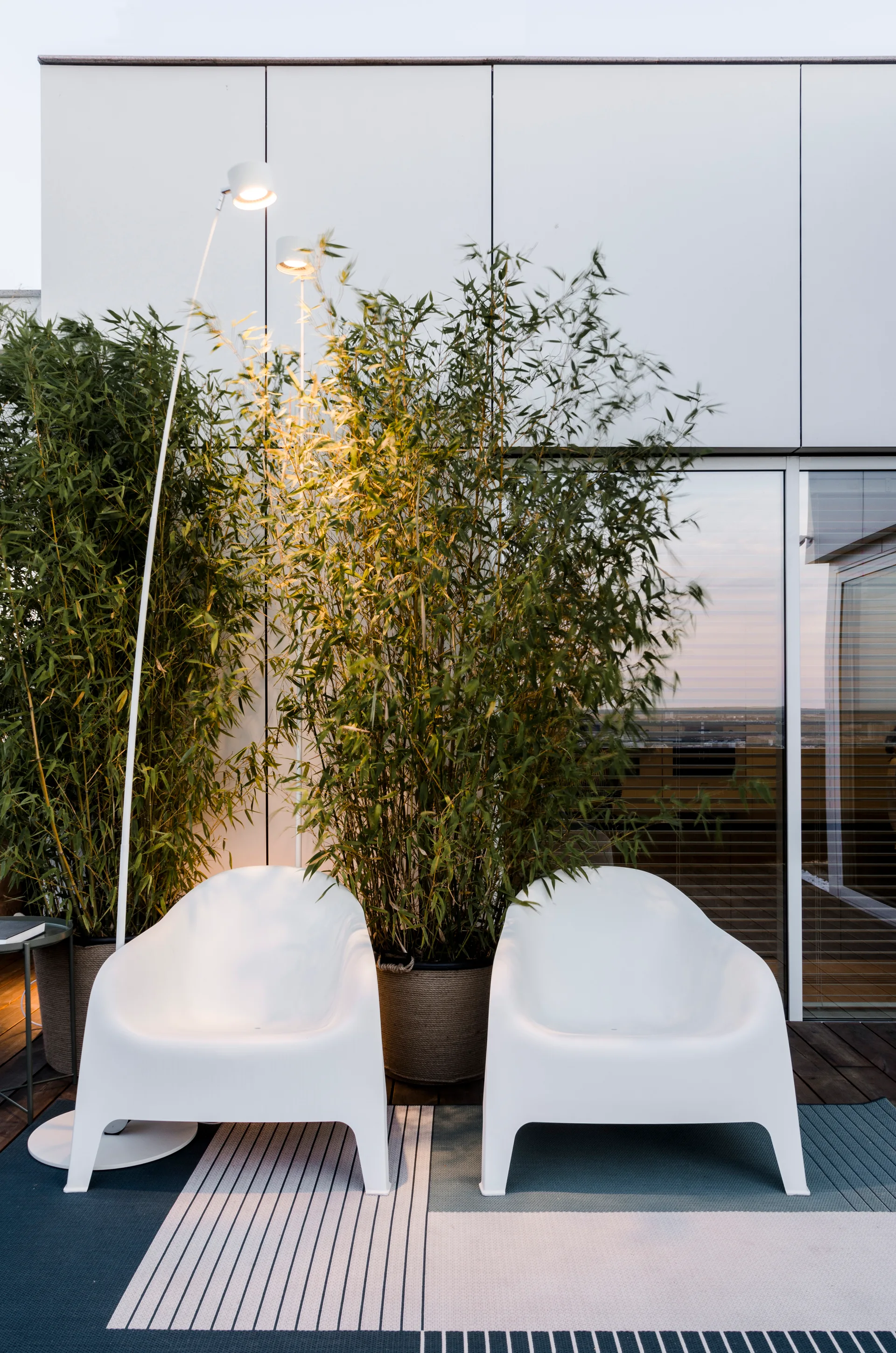 Two modern white armchairs sit on a striped rug next to tall potted bamboo plants on a wooden terrace deck.