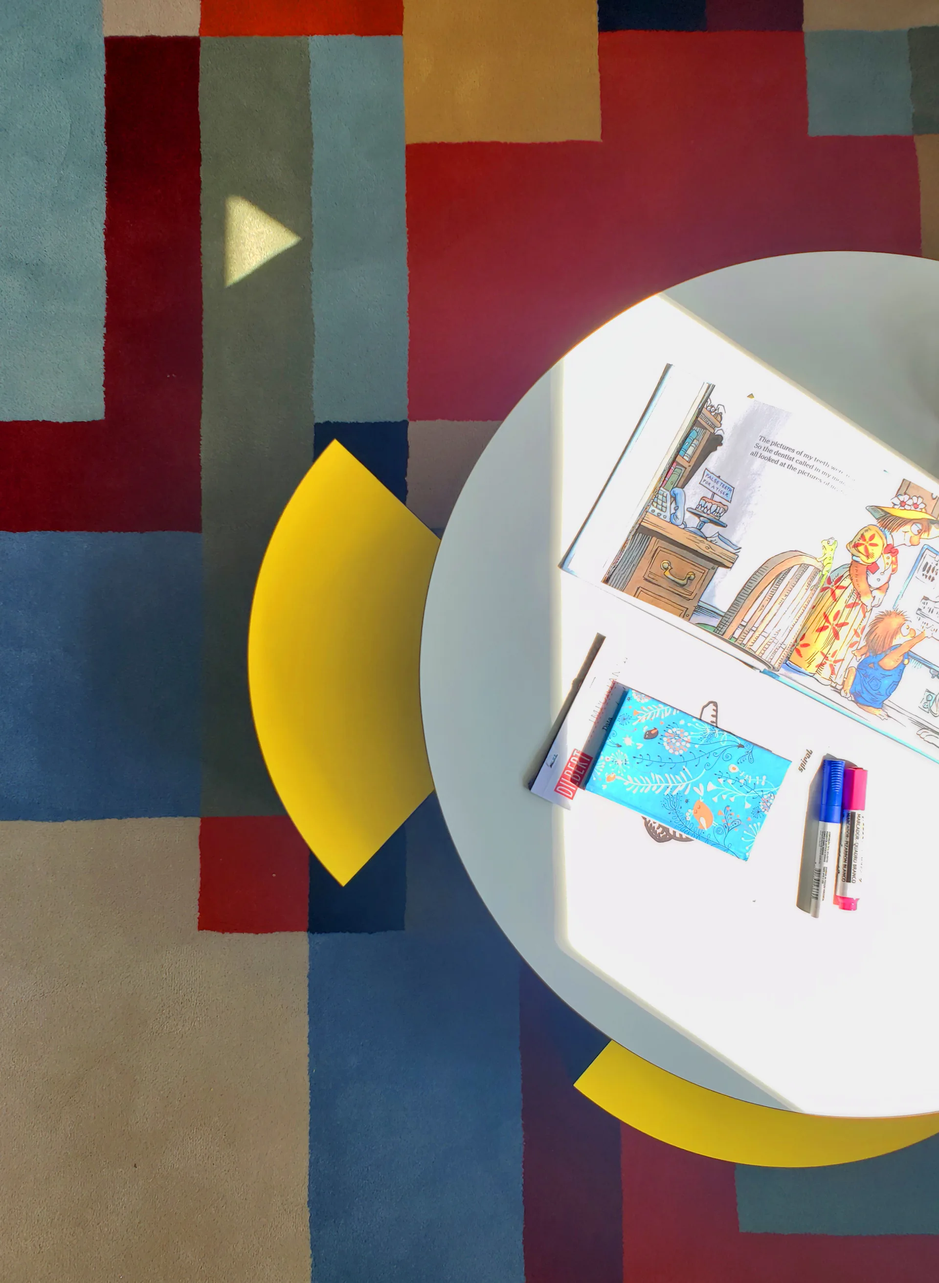 An overhead view of a round white coffee table and yellow chair on a vibrant red, blue, and yellow geometric block pattern area rug.