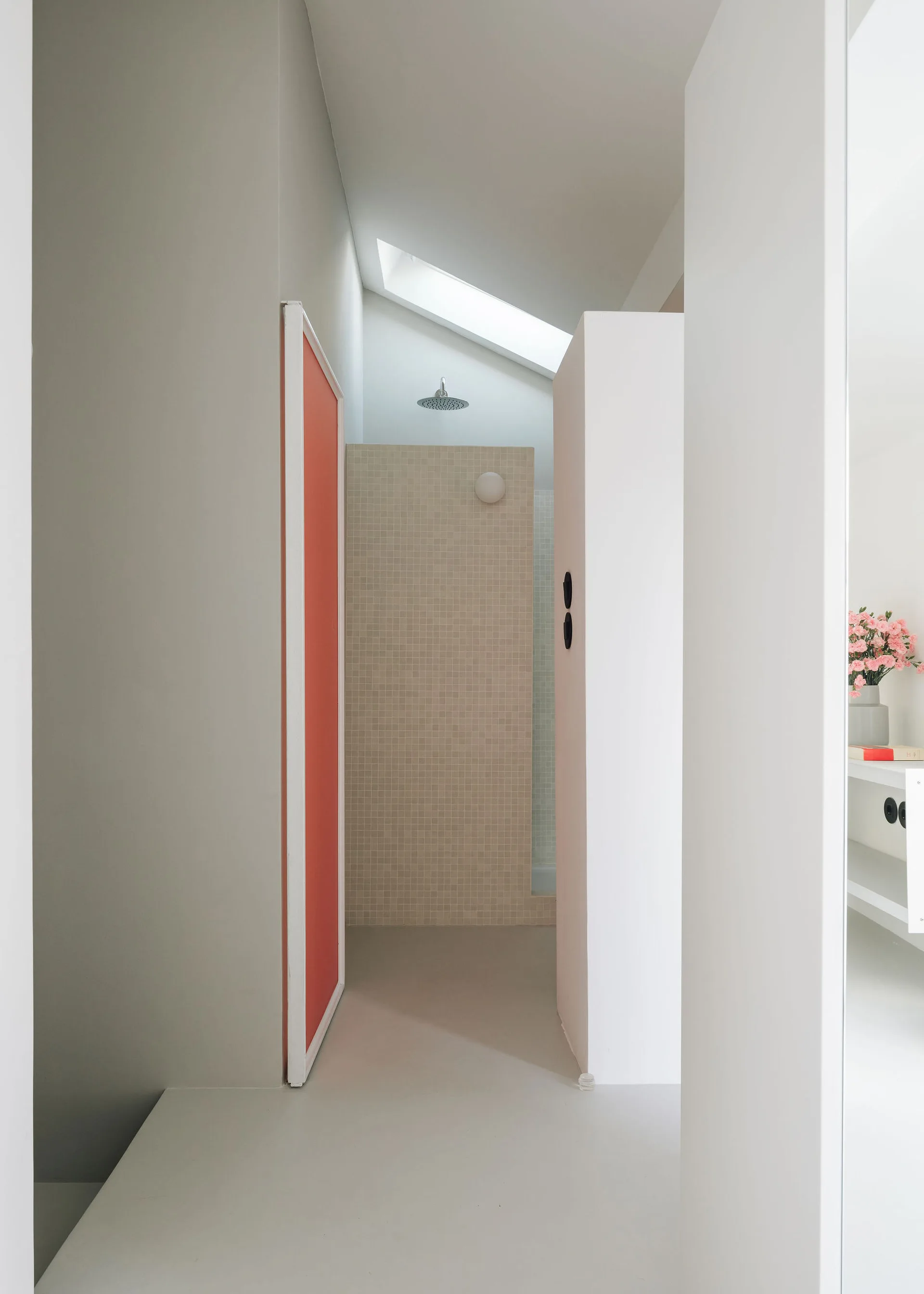 A minimalist bathroom entryway with an orange door panel, white partition walls, and a beige tiled shower under a skylight.