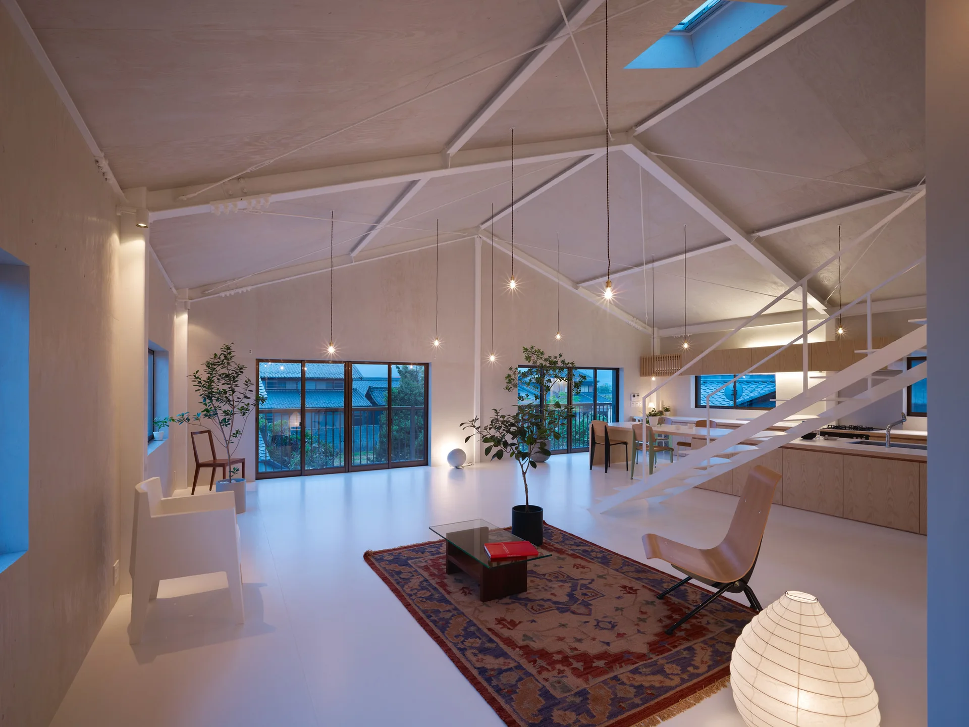 A spacious open-concept room features white floors, a vaulted plywood ceiling with white steel rafters, hanging bulb lights, and a patterned rug.