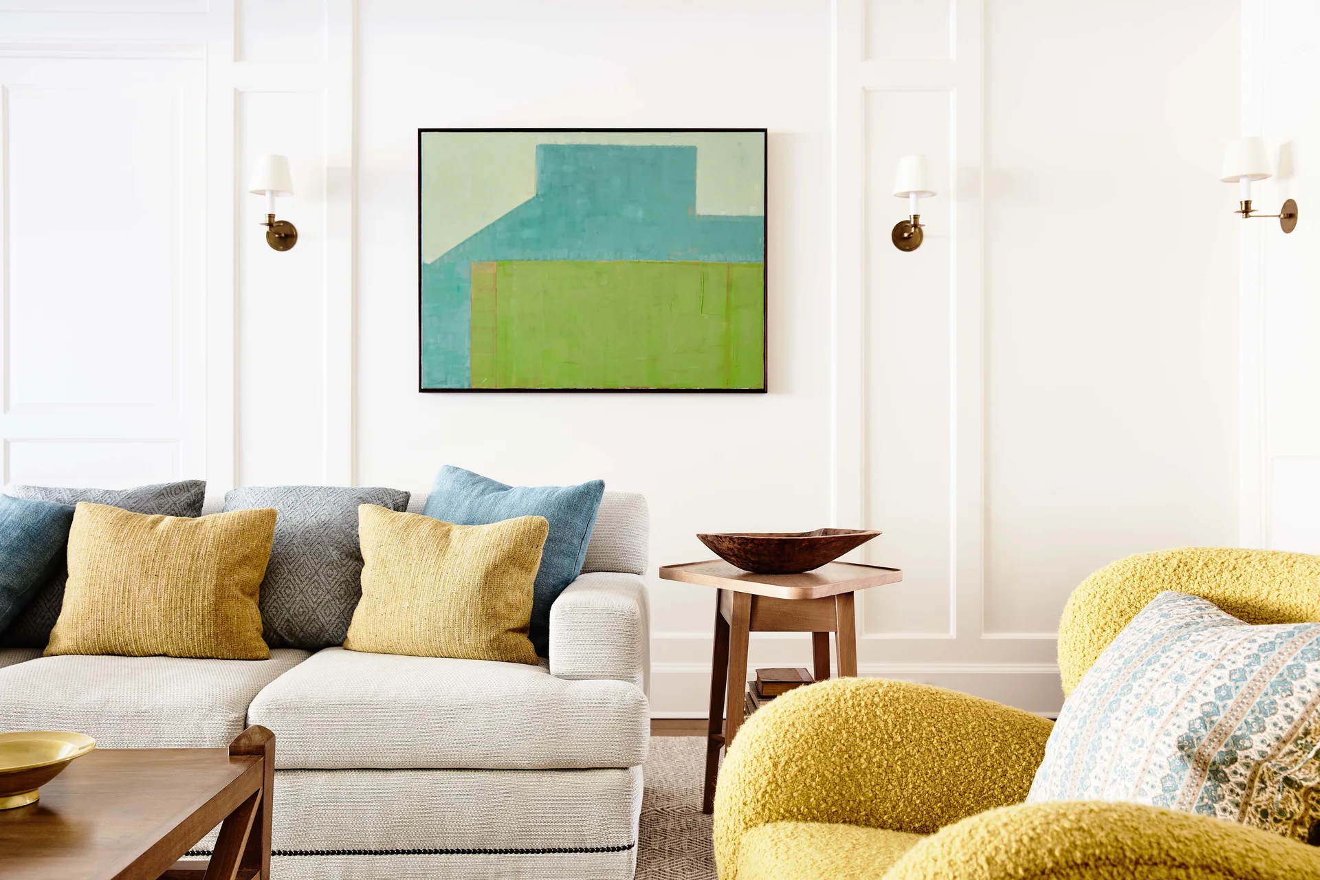 A light gray sofa with yellow and blue pillows sits next to a textured yellow armchair in a living room featuring white wood wall molding.