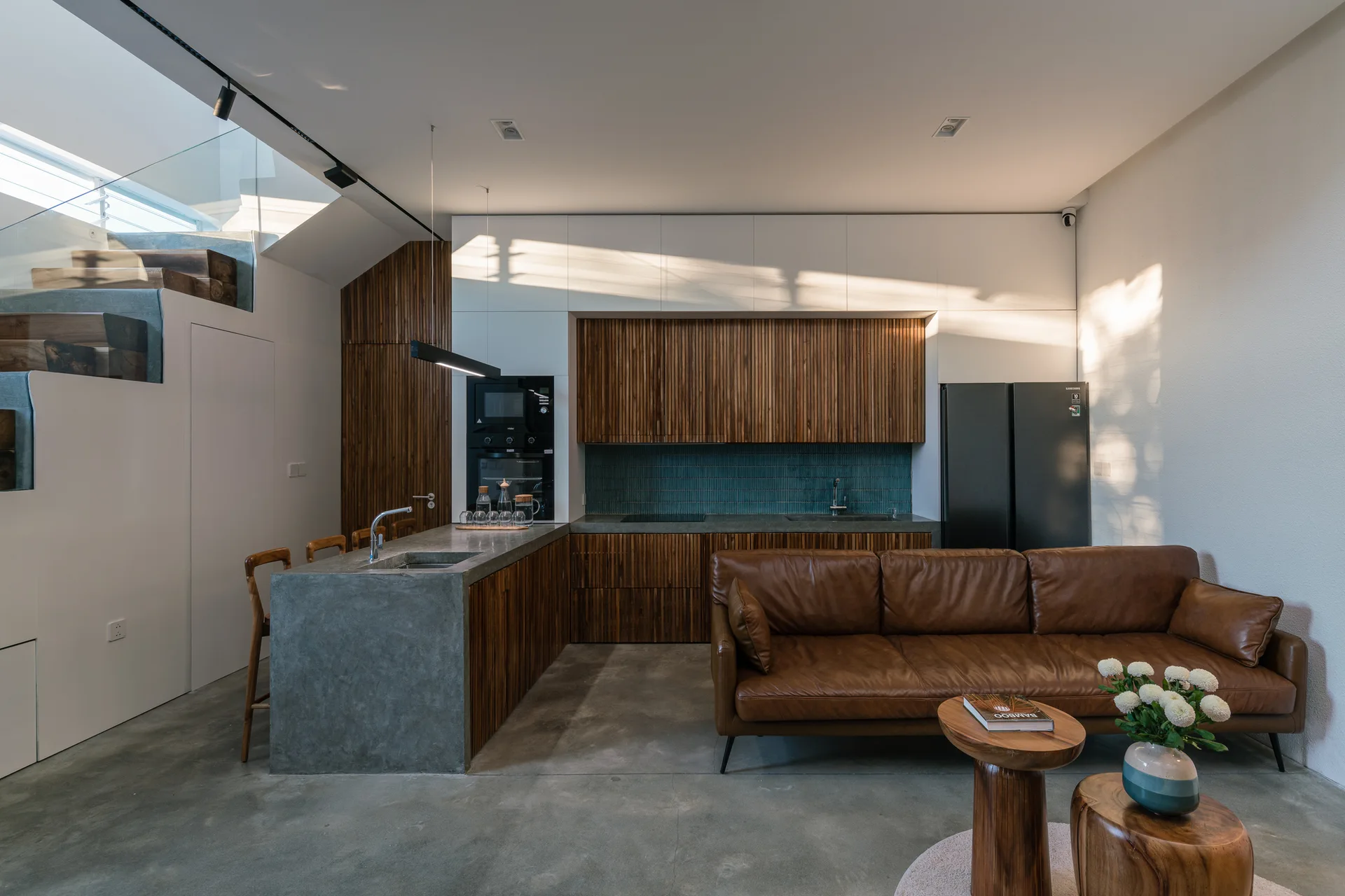 A modern open-plan kitchen features slatted wood cabinetry, a concrete island, and a brown leather sofa on a polished concrete floor.
