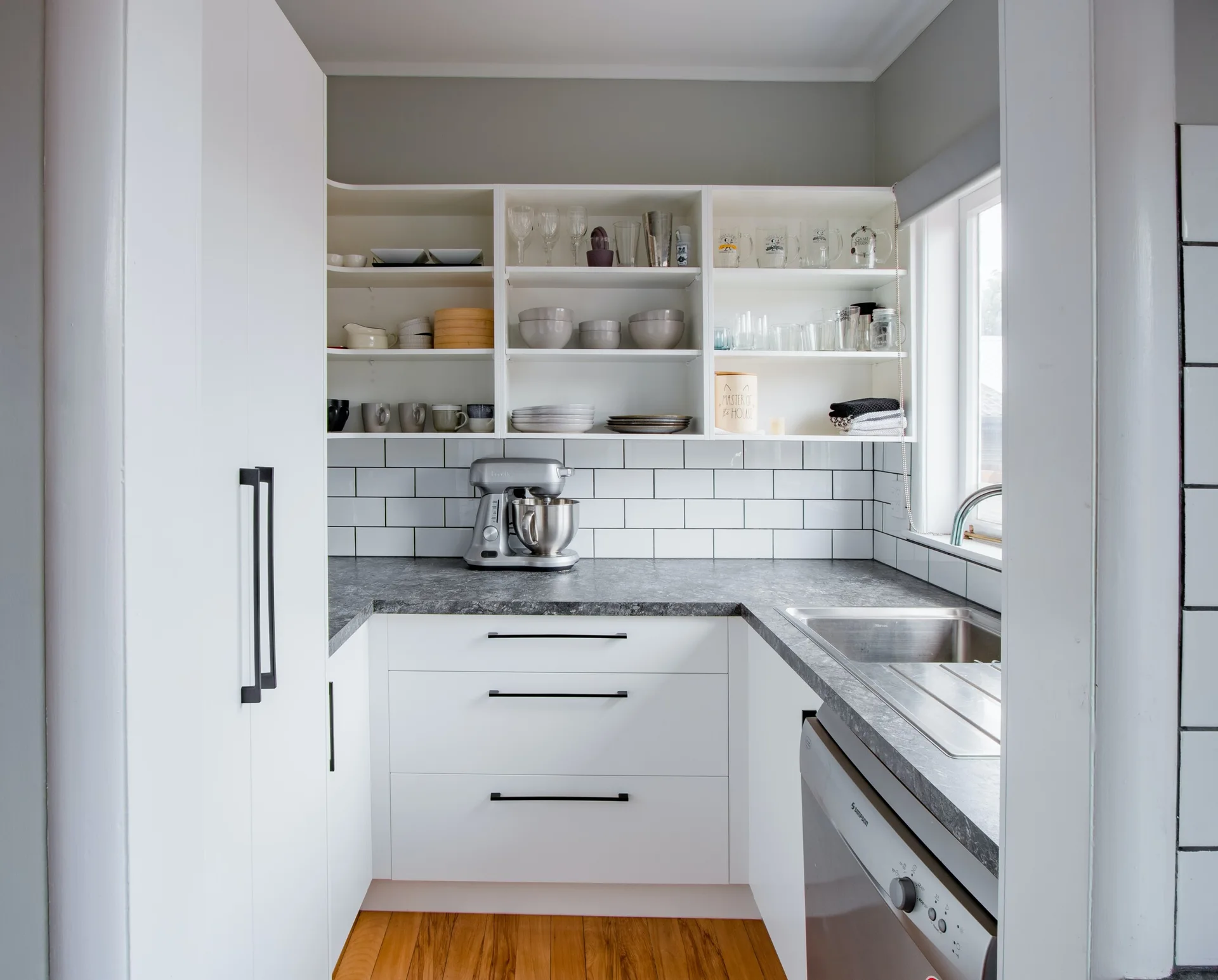 A compact U-shaped kitchen features white cabinets with black handles, gray countertops, a white subway tile backsplash, and three tiers of open shelving.