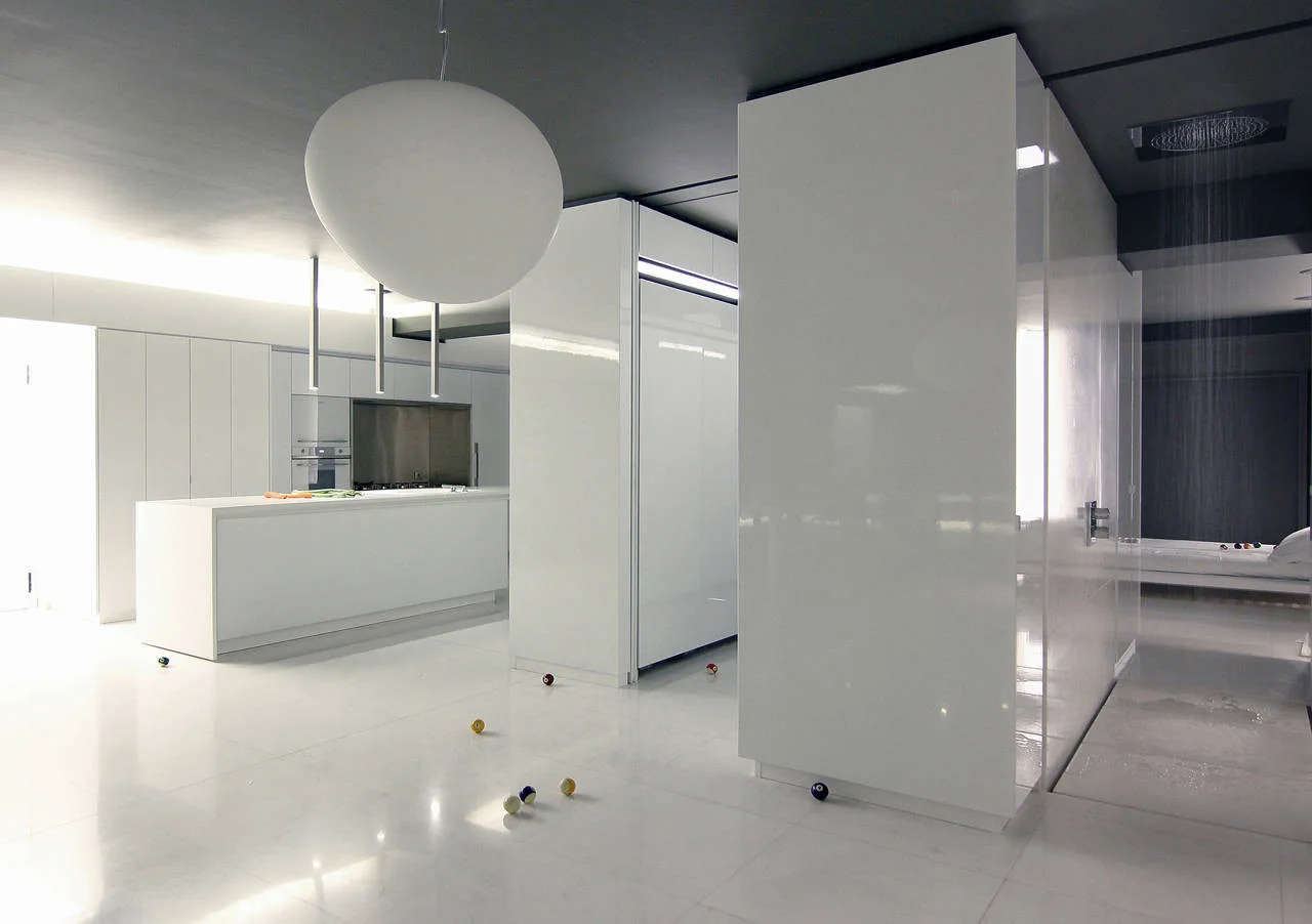 A minimalist kitchen features glossy white cabinetry, a large white island, and a dark grey ceiling with an overhead rain shower in the background.