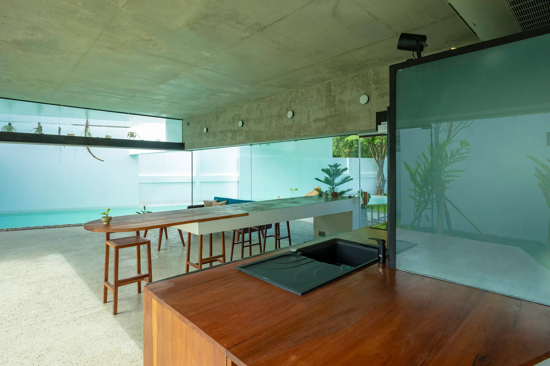 A minimalist kitchen featuring a wood island countertop, raw concrete ceiling, and a large glass wall looking out to a blue swimming pool.