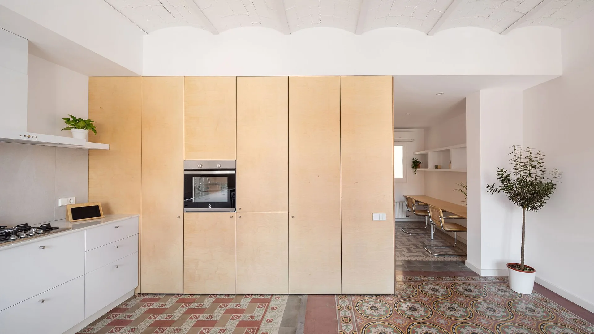 A modern kitchen with a full-height wall of natural plywood cabinetry, white countertops, and decorative patterned tile flooring.