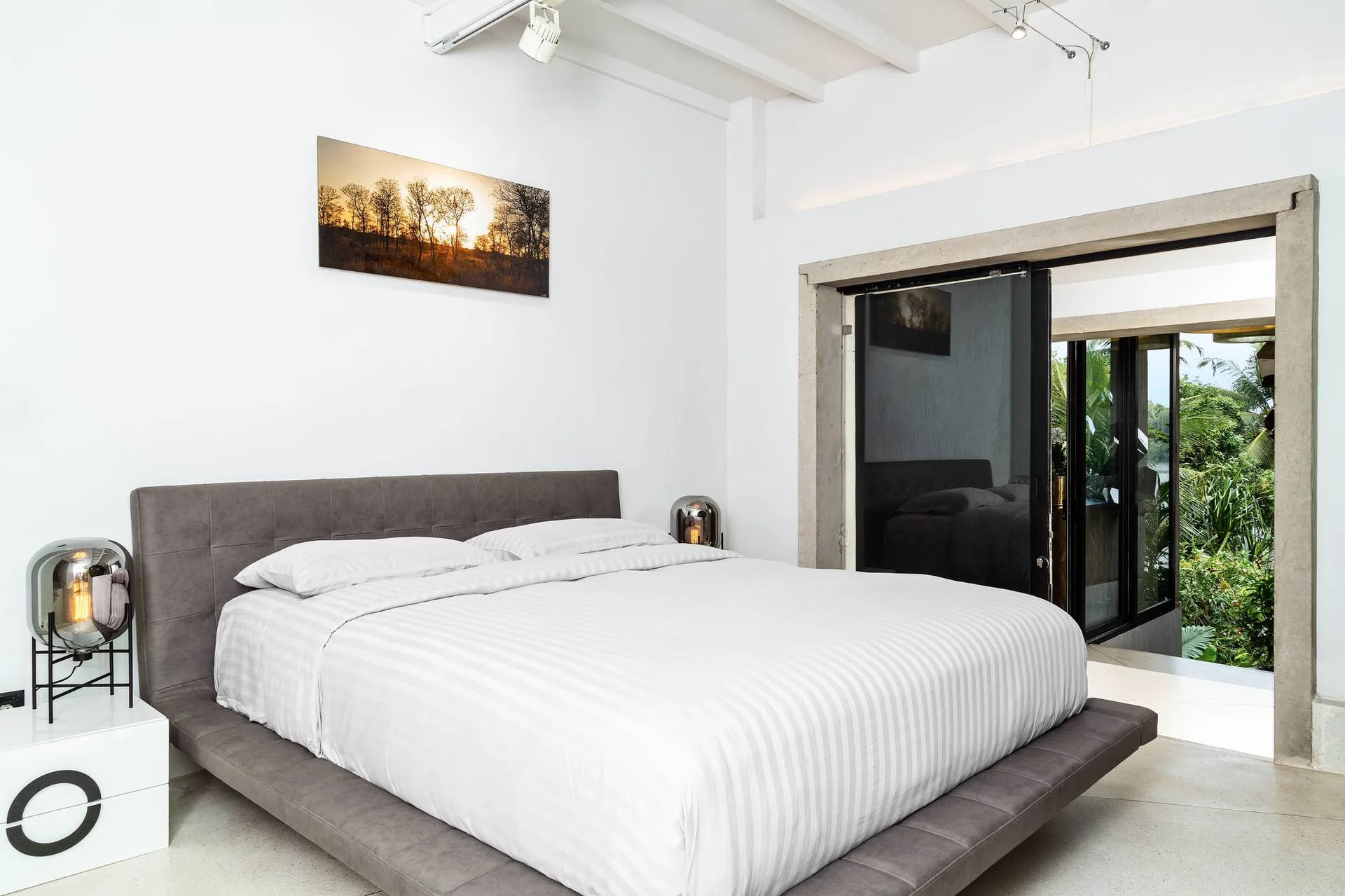 A minimalist bedroom features a low grey upholstered bed with striped white sheets next to a large sliding glass door with a concrete frame.