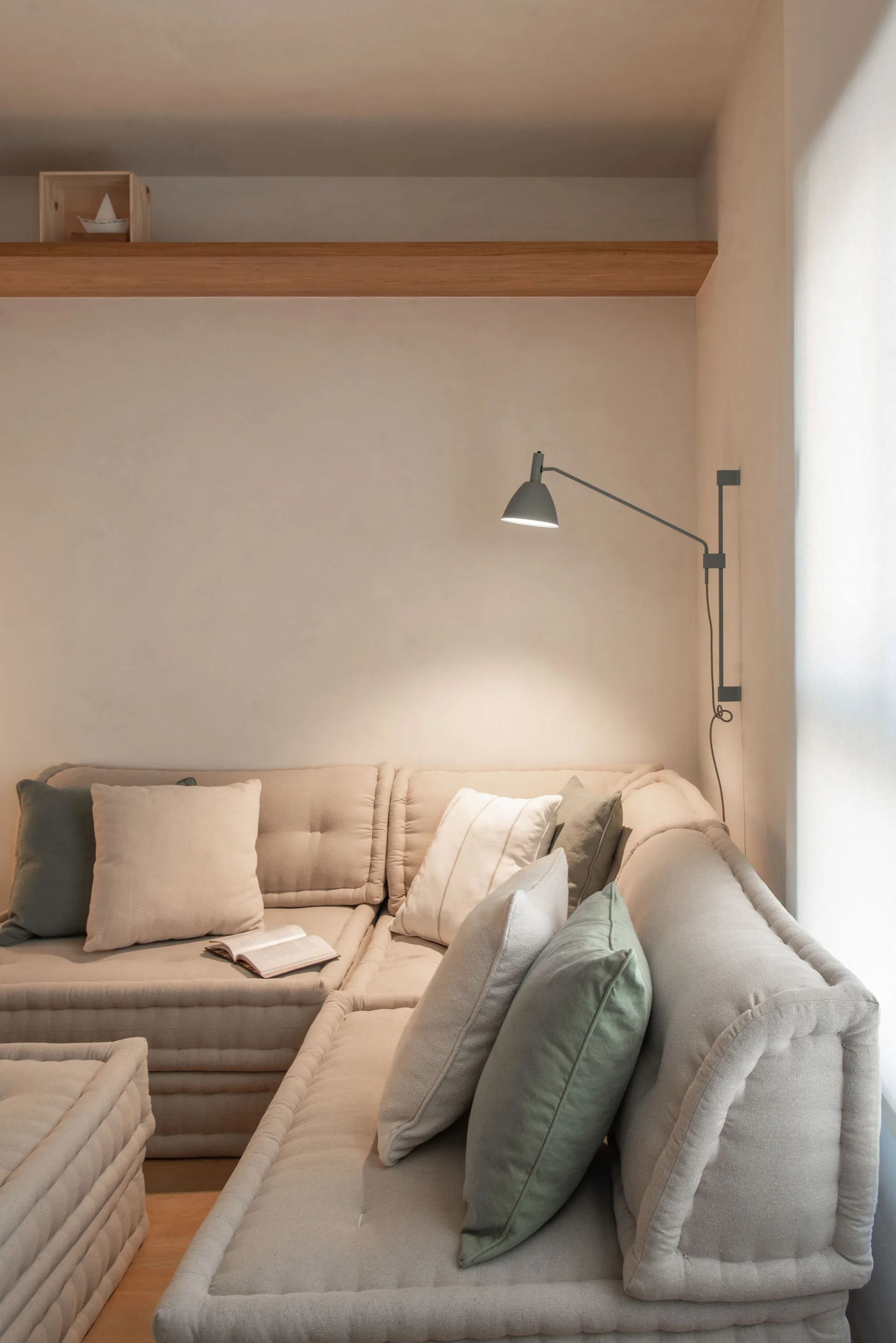 A beige modular corner sofa features light green and cream pillows beneath a floating wooden shelf and an adjustable grey wall lamp.