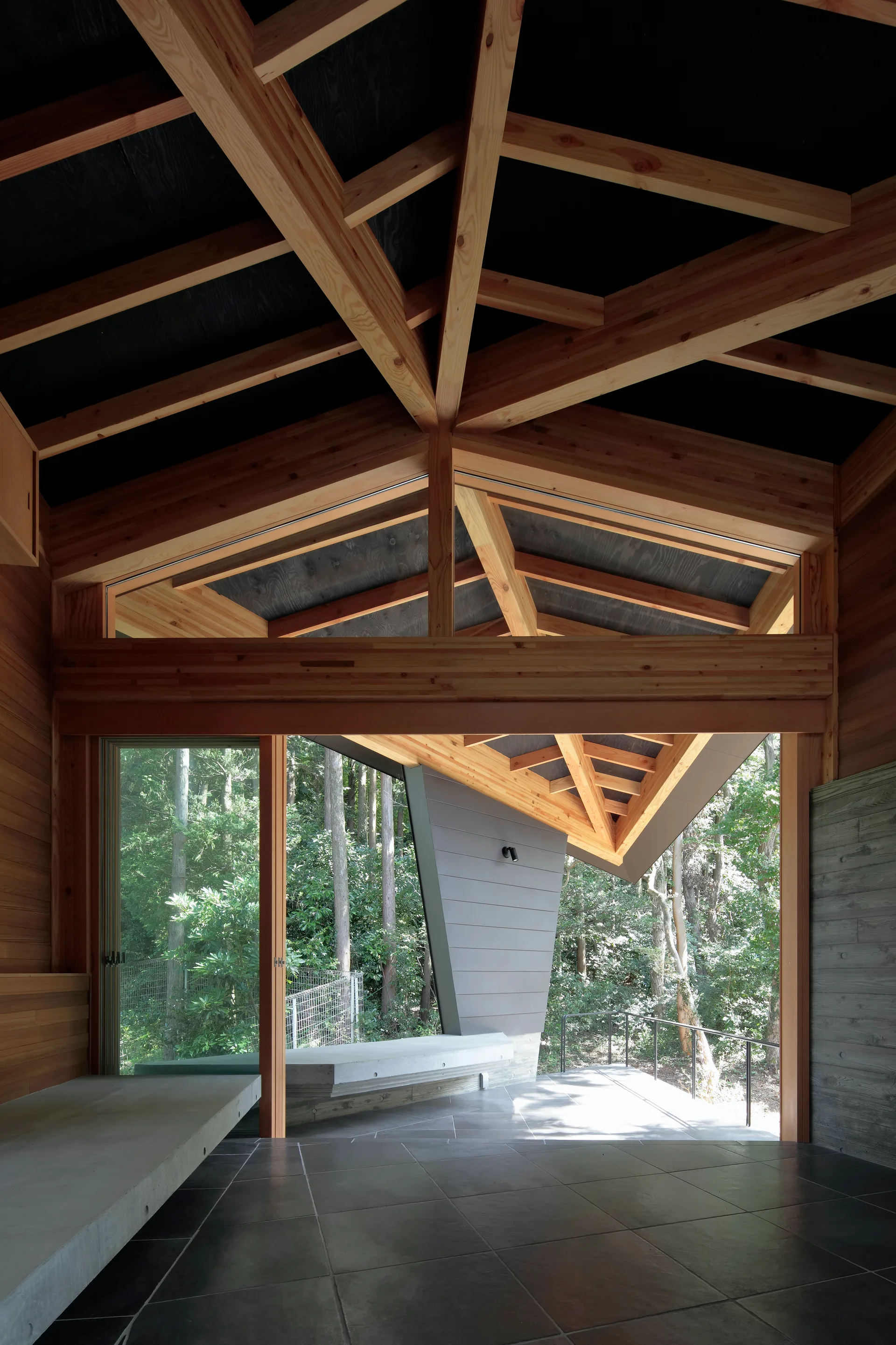 An interior space features complex geometric timber ceiling beams, large sliding glass doors opening to a forest, and a long concrete bench.
