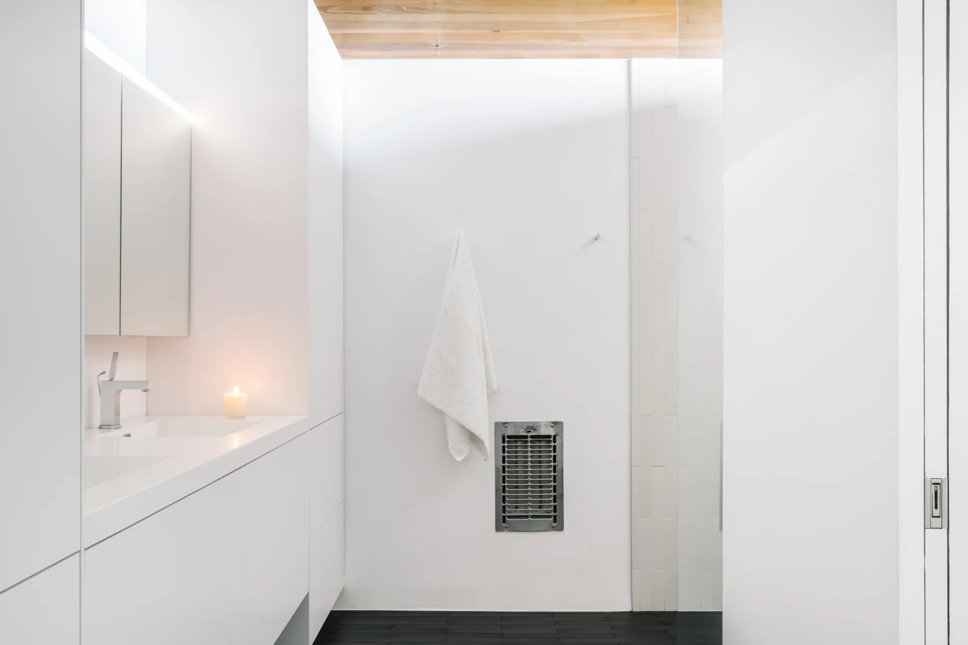 A minimalist white bathroom features a flat-front vanity with a chrome faucet, black tile flooring, and an exposed wood plank ceiling.