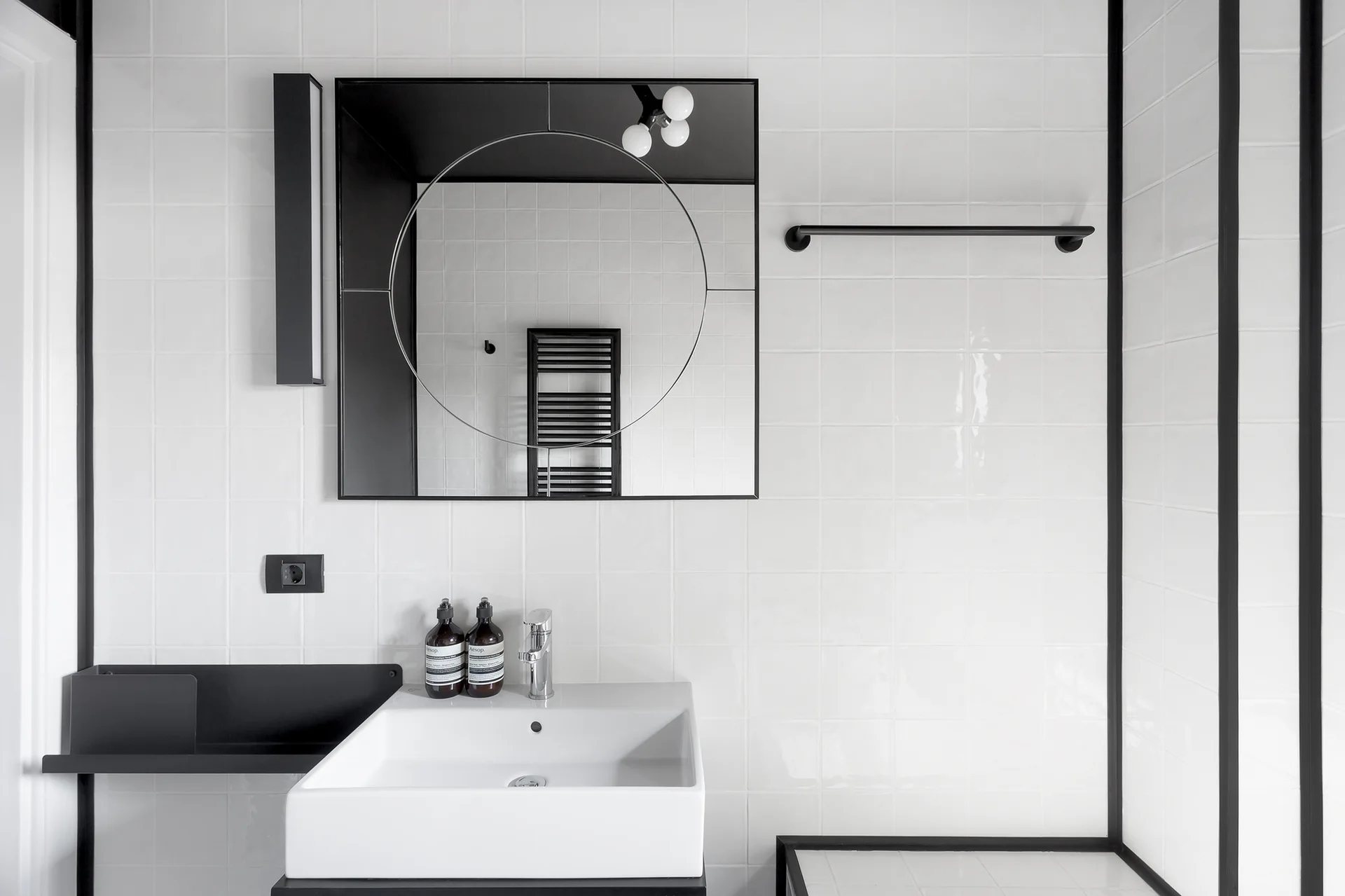 A modern bathroom features white square tiles, a white ceramic sink, matte black metal fixtures, and a rectangular mirror with a circular design.
