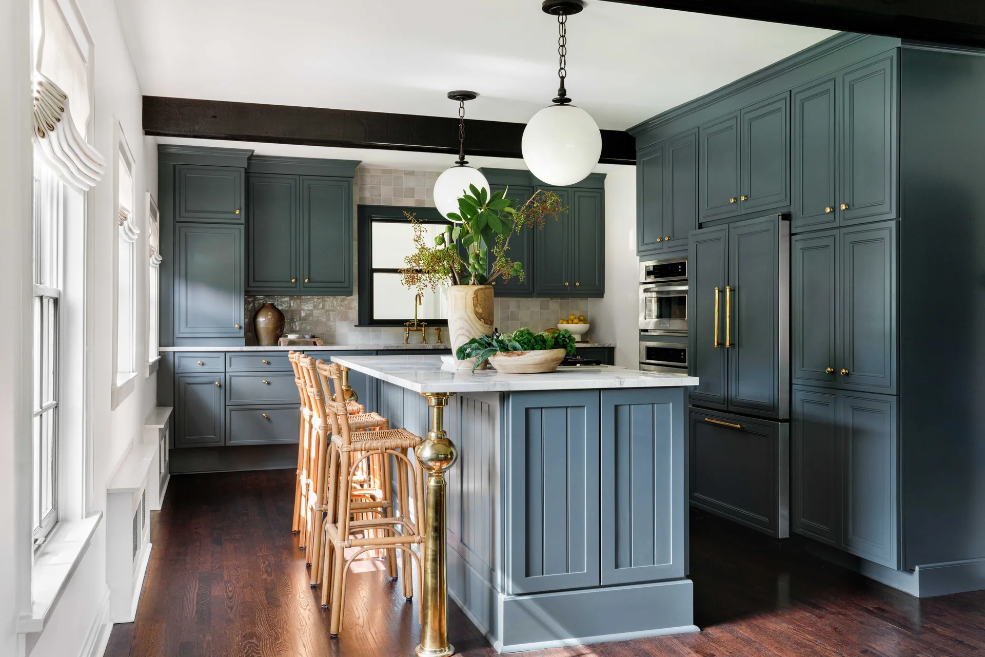 A transitional kitchen features dark grey-blue cabinetry, a marble-topped island with rattan bar stools, globe pendant lights, and dark wood floors.