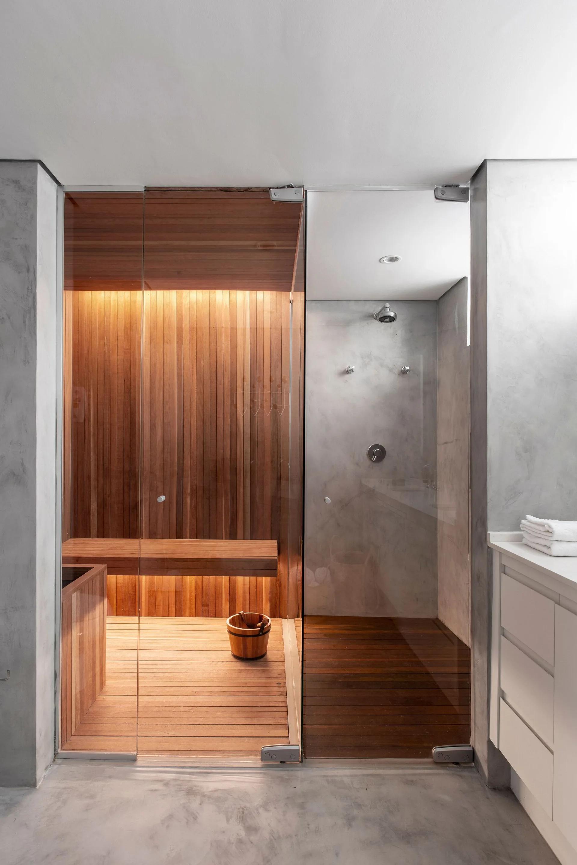 A glass-enclosed bathroom setup features a wood-paneled sauna on the left next to a concrete-walled shower stall on the right.