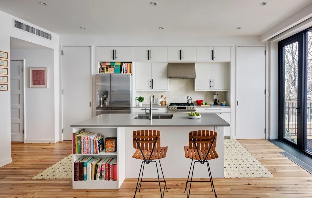A modern white kitchen features a central island with a grey countertop, built-in bookshelf, and two slatted wood bar stools.