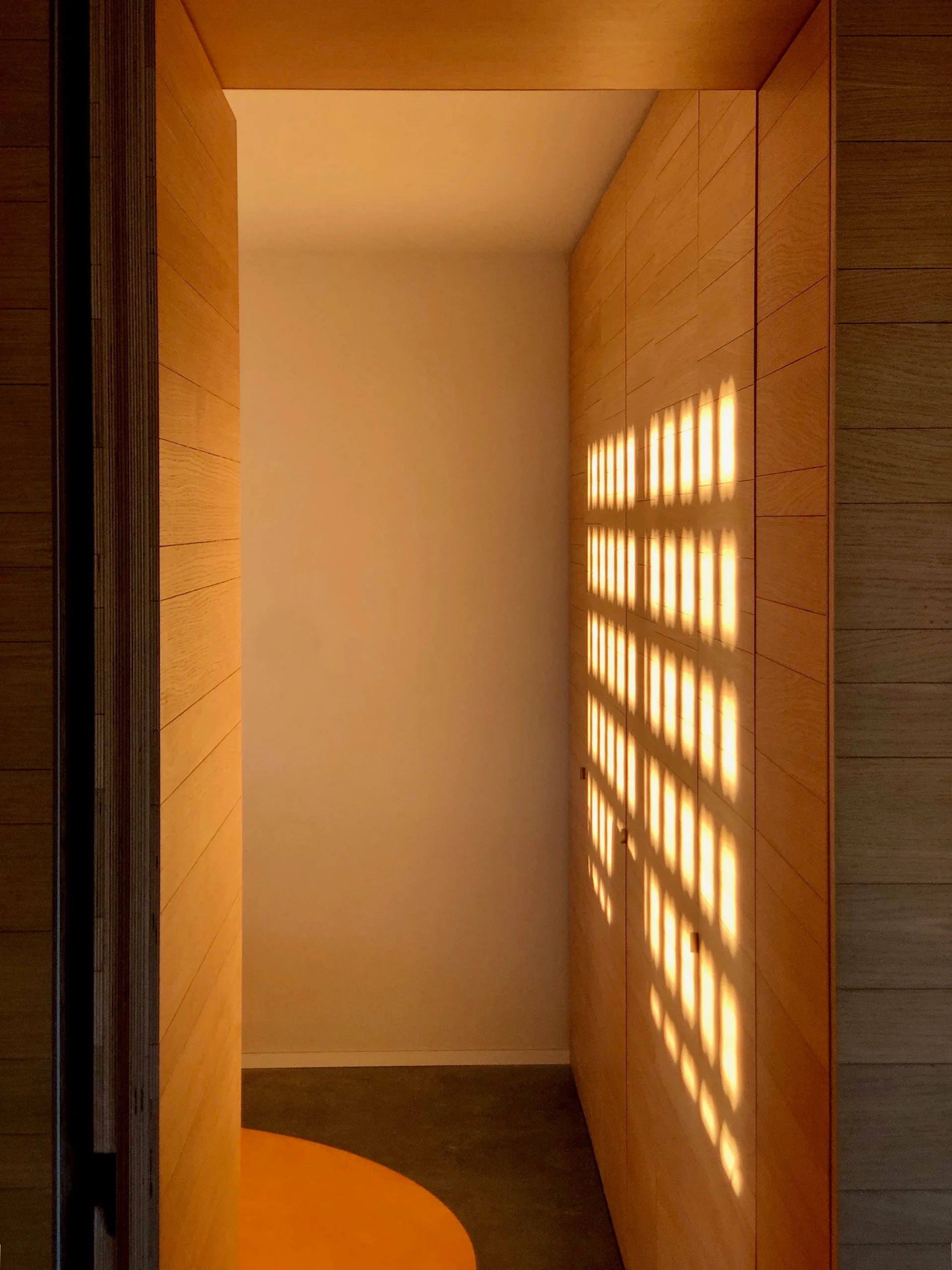 A minimalist hallway featuring horizontal natural wood paneling, concrete floors, and a geometric grid pattern of warm light projected onto the wall.