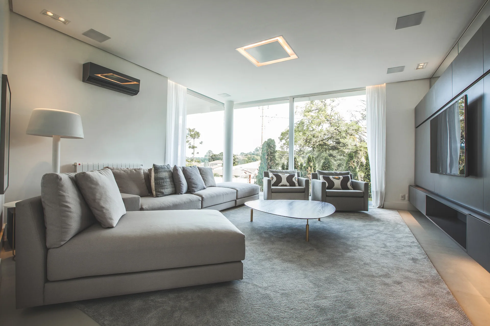 A minimalist grey living room features a large sectional sofa, two armchairs, an oval wood coffee table, and expansive floor-to-ceiling windows.