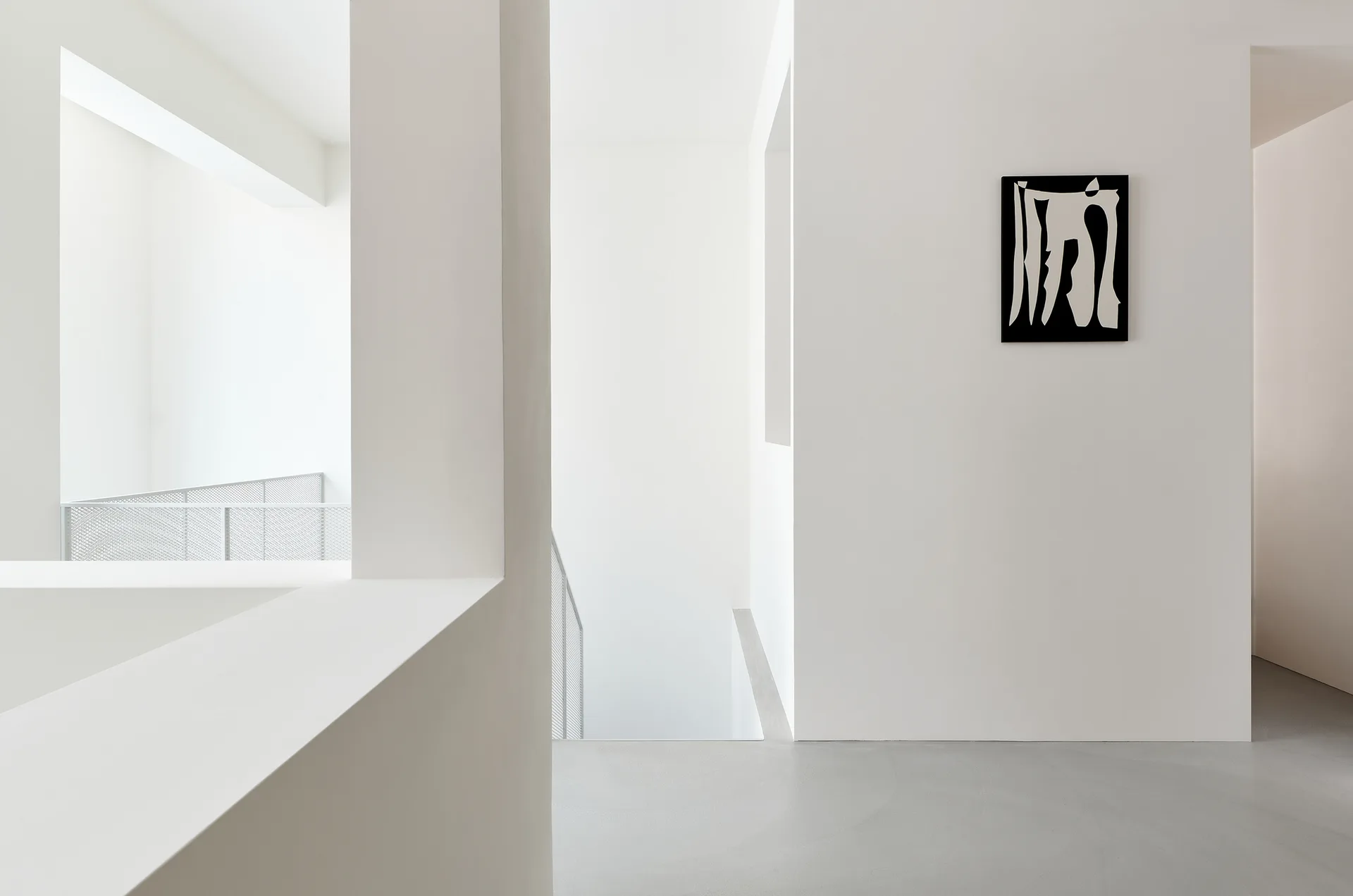 Minimalist white hallway with a gray floor, mesh staircase railing, sharp geometric wall cutouts, and a small black and white framed artwork.