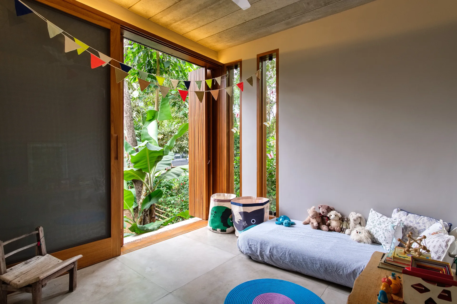 A bright kids' bedroom features a low floor bed, colorful bunting, and large wooden sliding glass doors that open to a lush green garden.