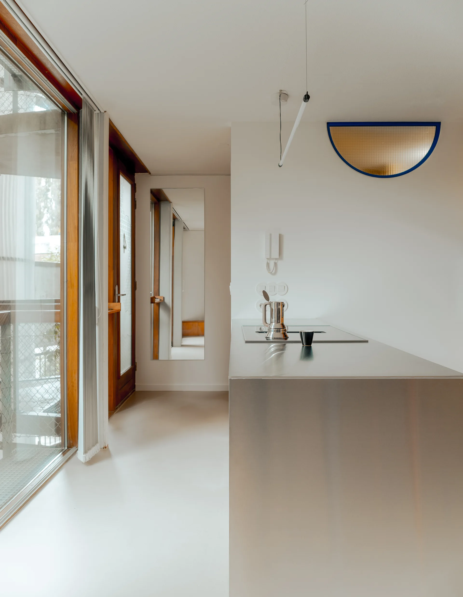 A modern minimalist kitchen featuring a stainless steel island next to large wood-framed glass doors and a white wall with an amber semi-circular sconce.