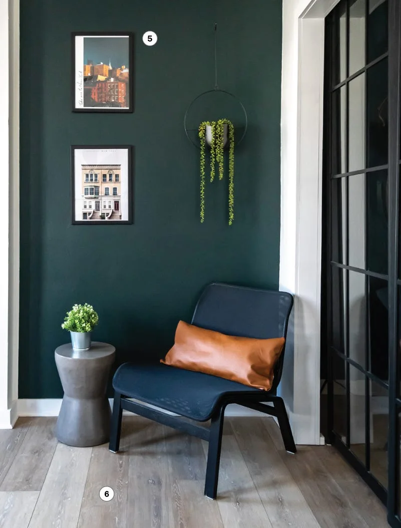 A modern corner features a dark green accent wall, a black accent chair with a tan lumbar pillow, and stacked framed wall art.