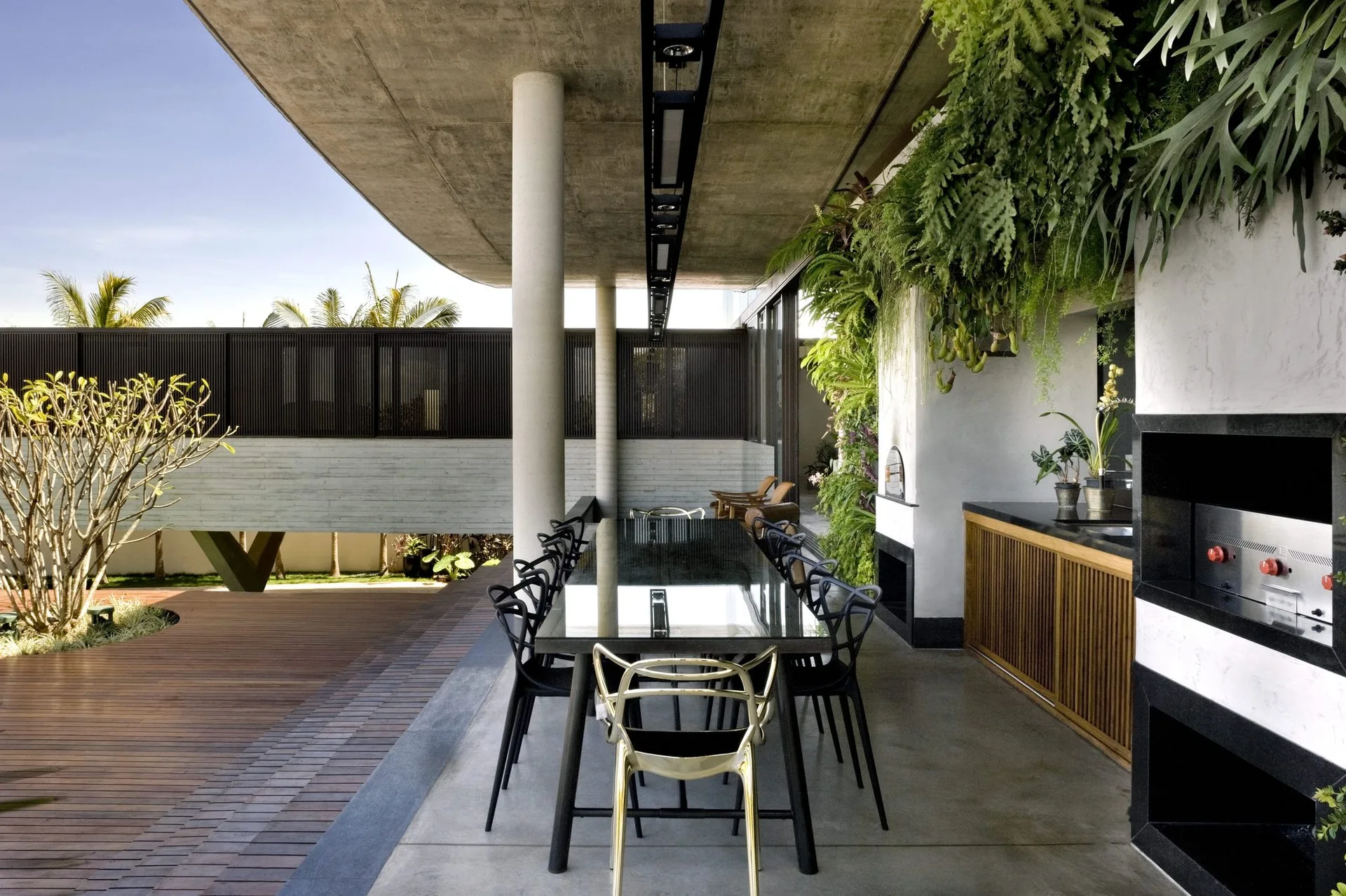 A covered concrete patio features a long black dining table, black chairs, a gold accent chair, an outdoor kitchen, and a lush vertical garden.
