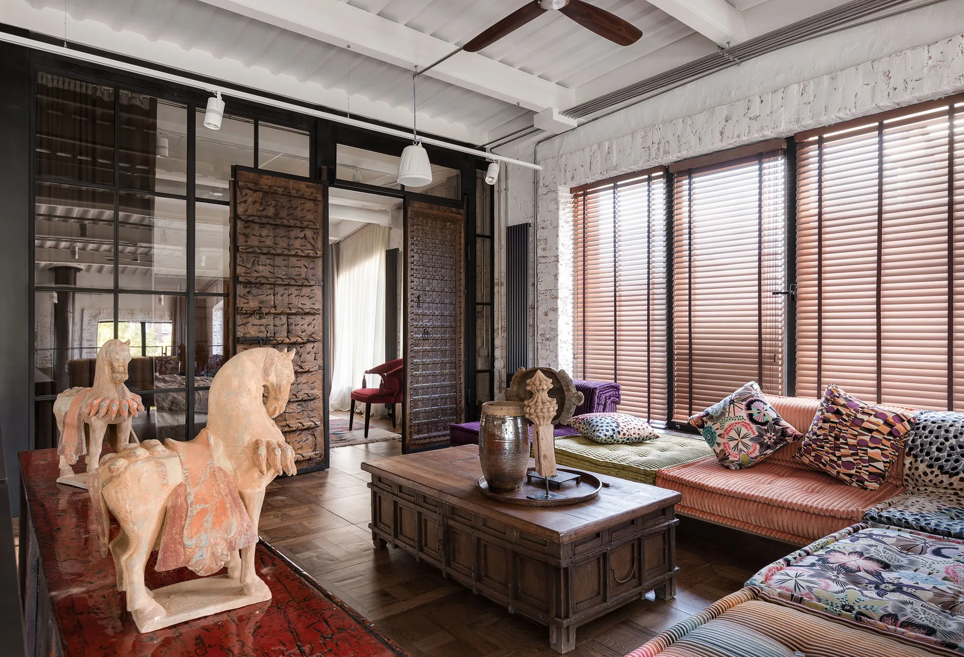 An eclectic living room featuring colorful low modular seating, a carved wooden coffee table, clay horse sculptures, and antique wooden partition doors.
