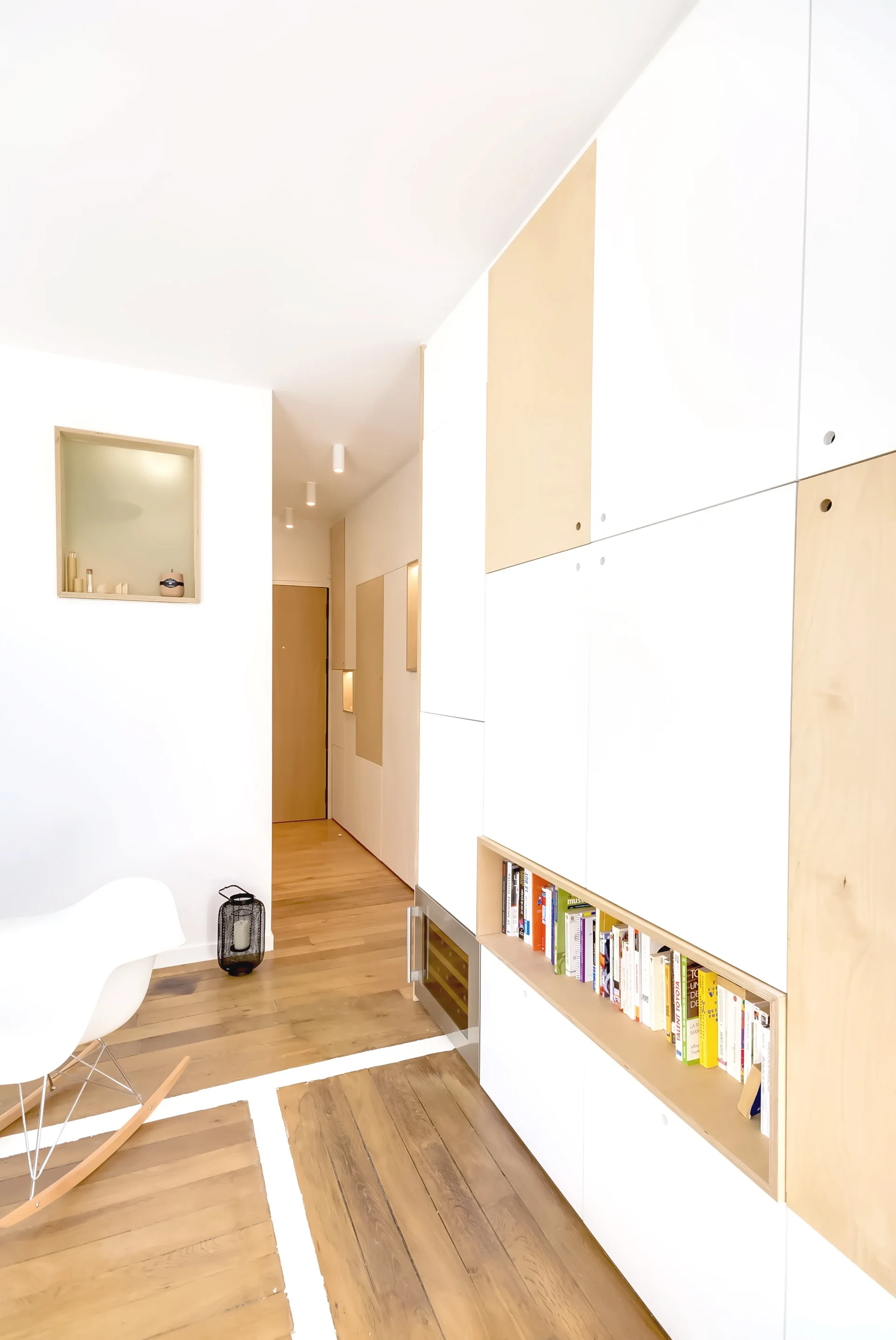Modern hallway featuring custom white and wood-toned cabinetry with a recessed bookshelf, wood flooring, and a white rocking chair.