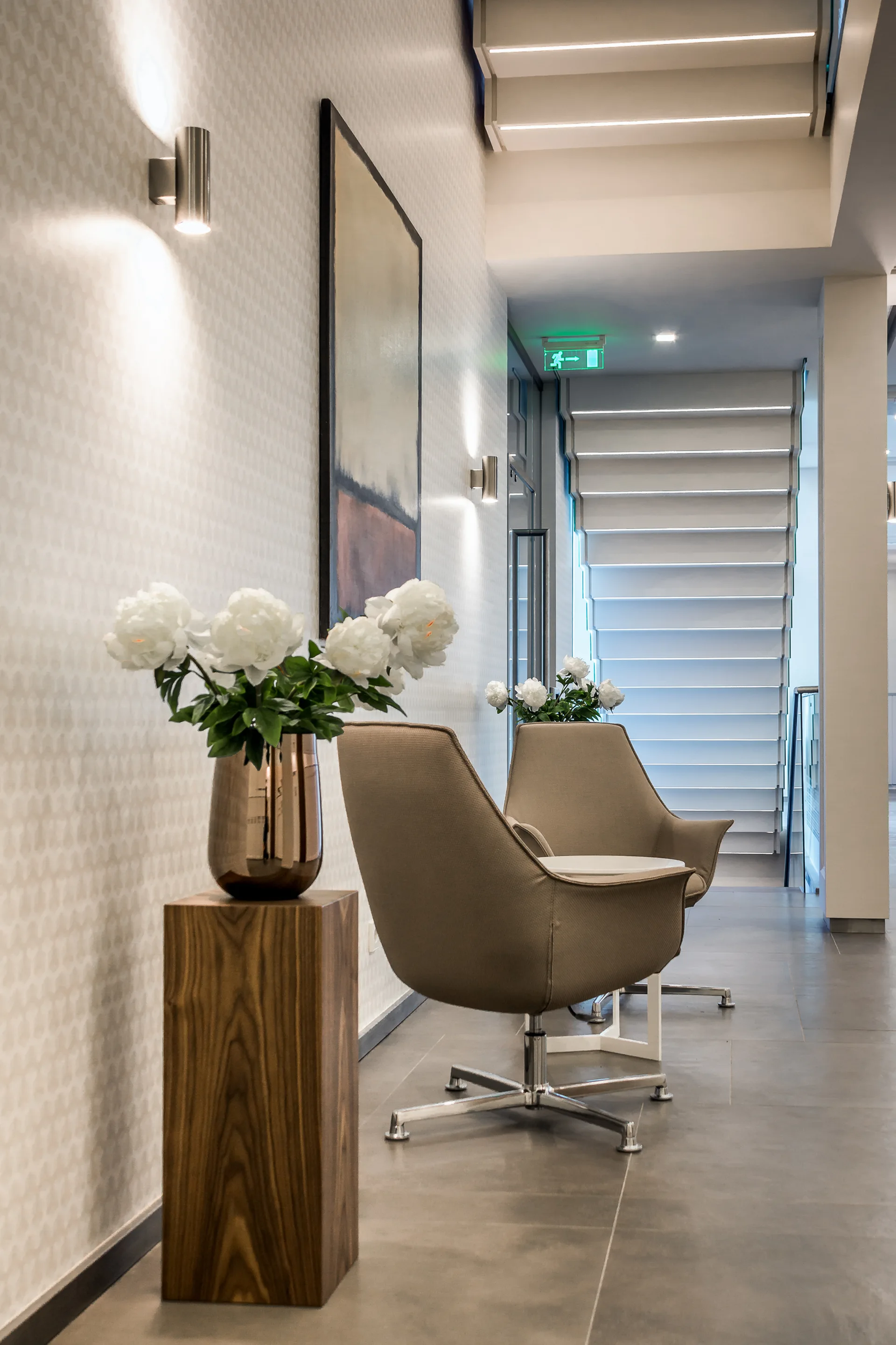 Two taupe armchairs sit next to a wooden pedestal with white flowers in a contemporary hallway featuring subtle patterned wallpaper.