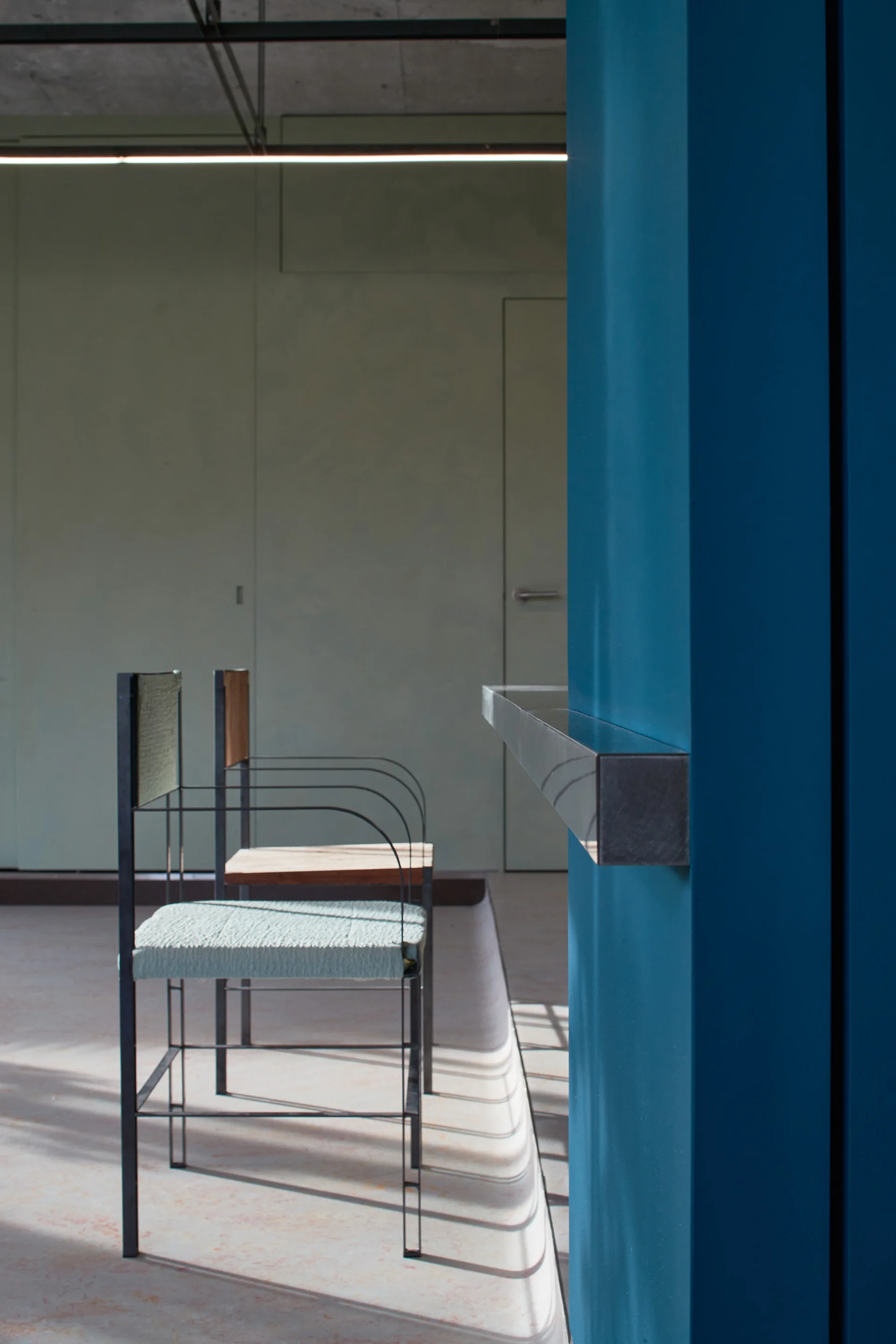 Two minimalist chairs with thin metal frames stand near a bright blue structural column under an exposed concrete ceiling.