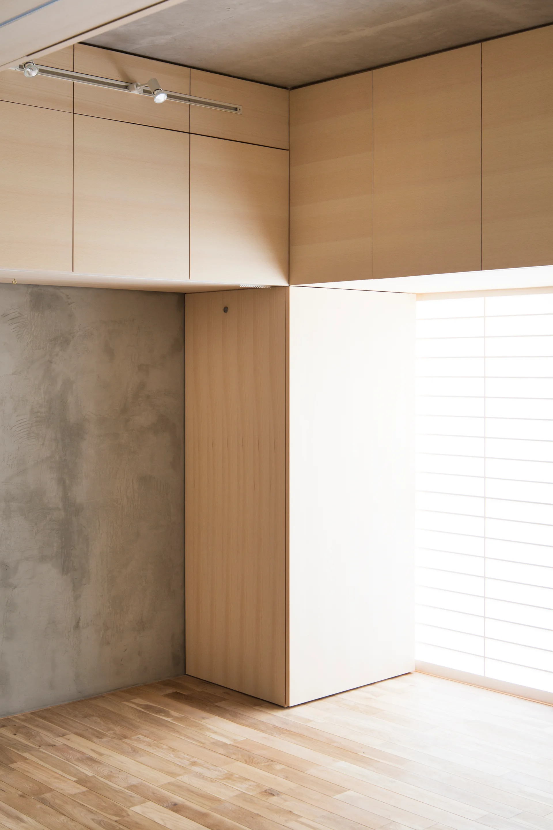 Minimalist light wood cabinets integrated next to a textured grey concrete wall and a gridded translucent window above light wood floors.