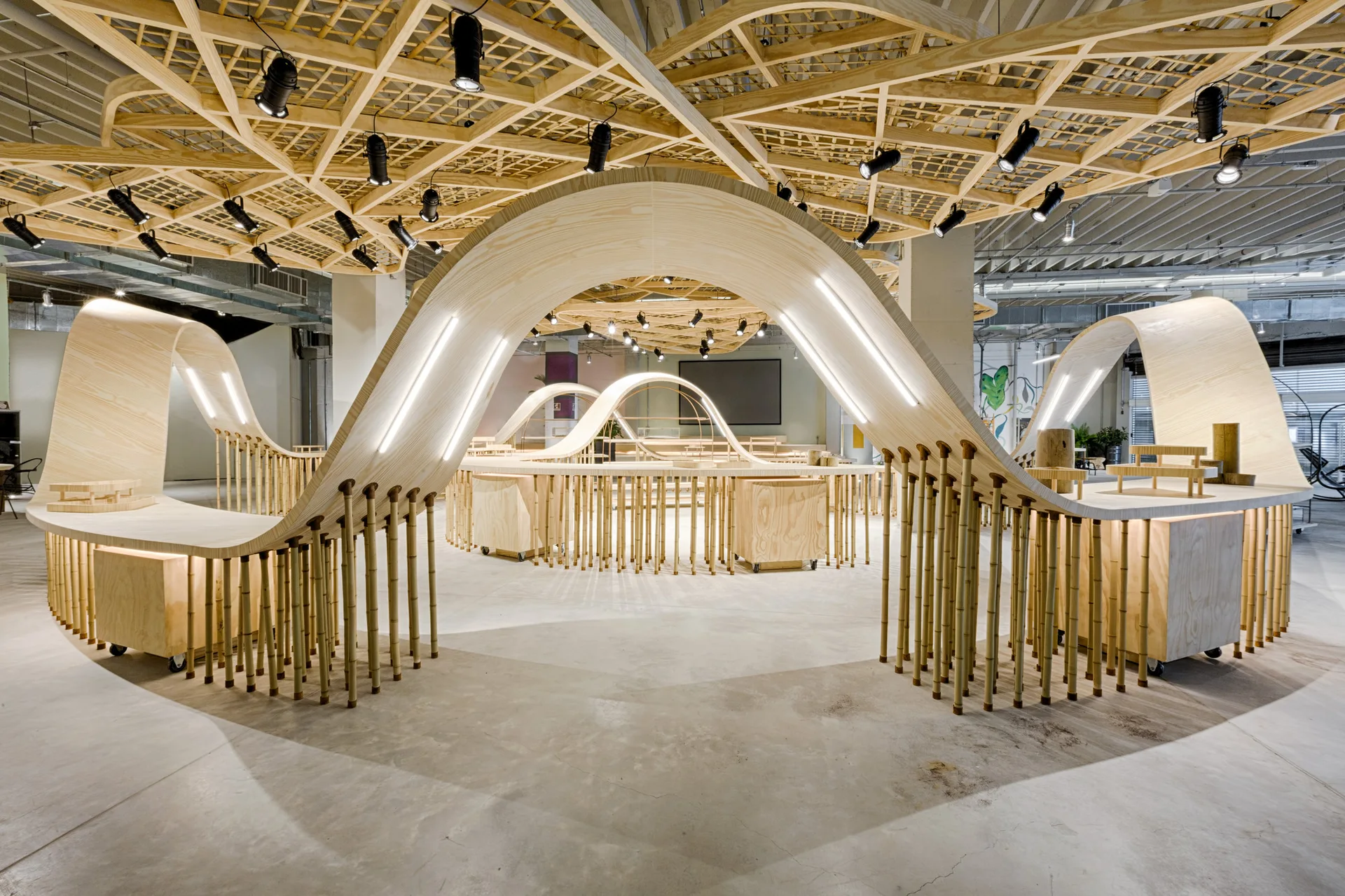 A commercial exhibition space features curving blonde wood display structures supported by vertical bamboo poles beneath a geometric wooden lattice ceiling.