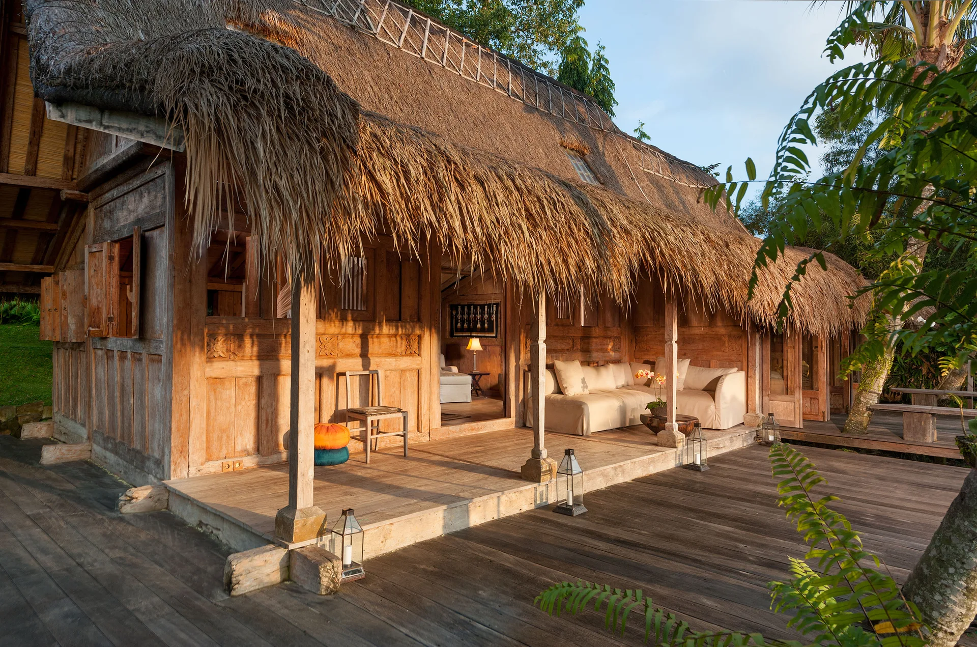 A rustic wooden porch with a prominent thatched roof features a cream-colored sectional sofa, metal floor lanterns, and carved wall panels.
