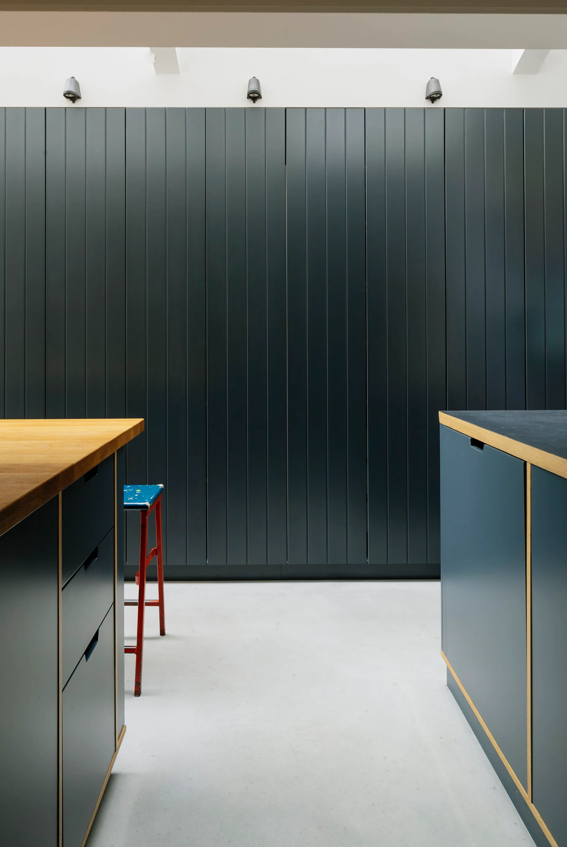 A modern kitchen with dark blue cabinetry featuring light wood trim, a vertical grooved wall cabinet, a red stool, and grey concrete flooring.