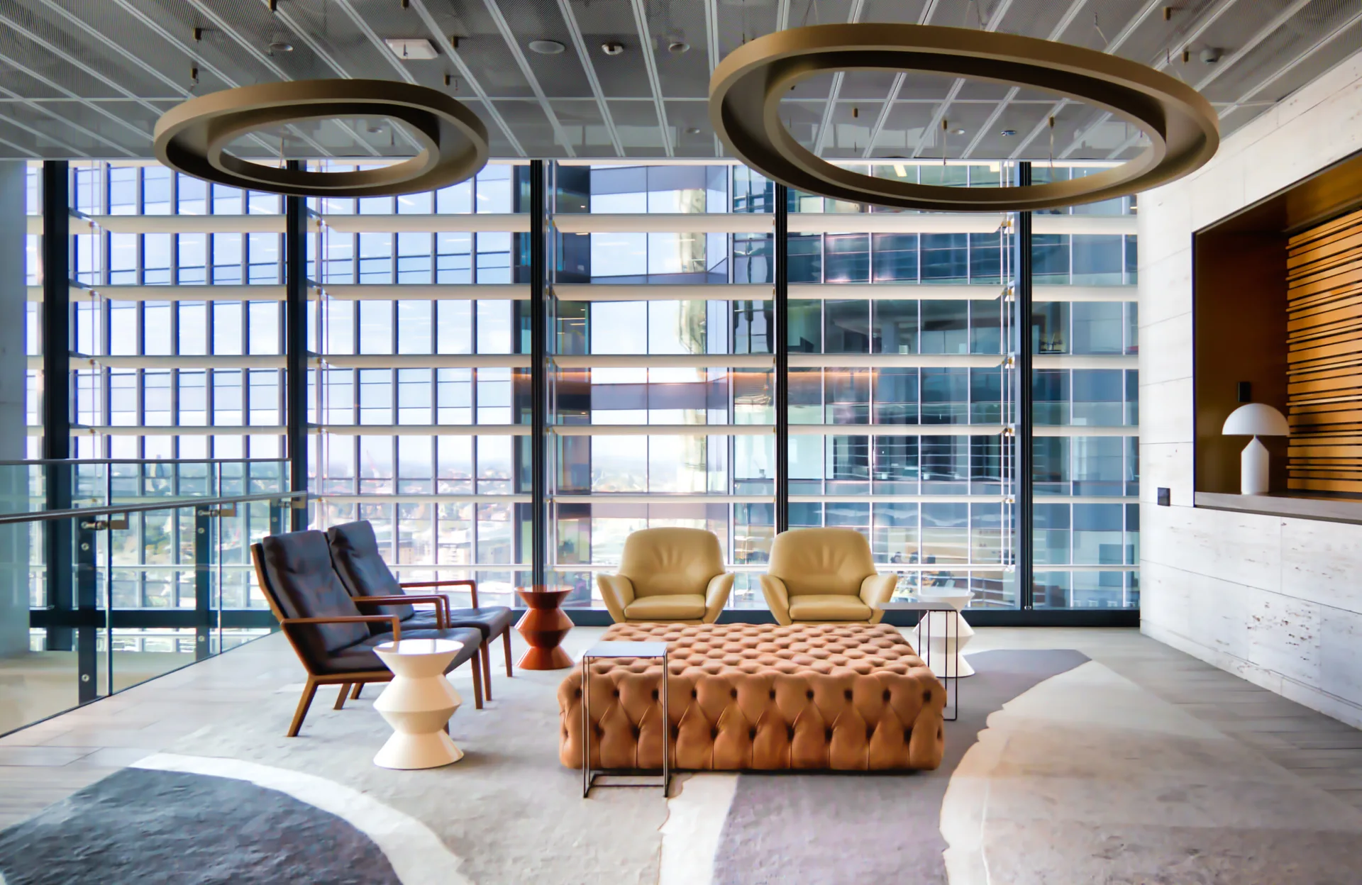 A modern lounge area features a large tan tufted ottoman, leather armchairs, and circular ring pendant lights against a massive window wall.