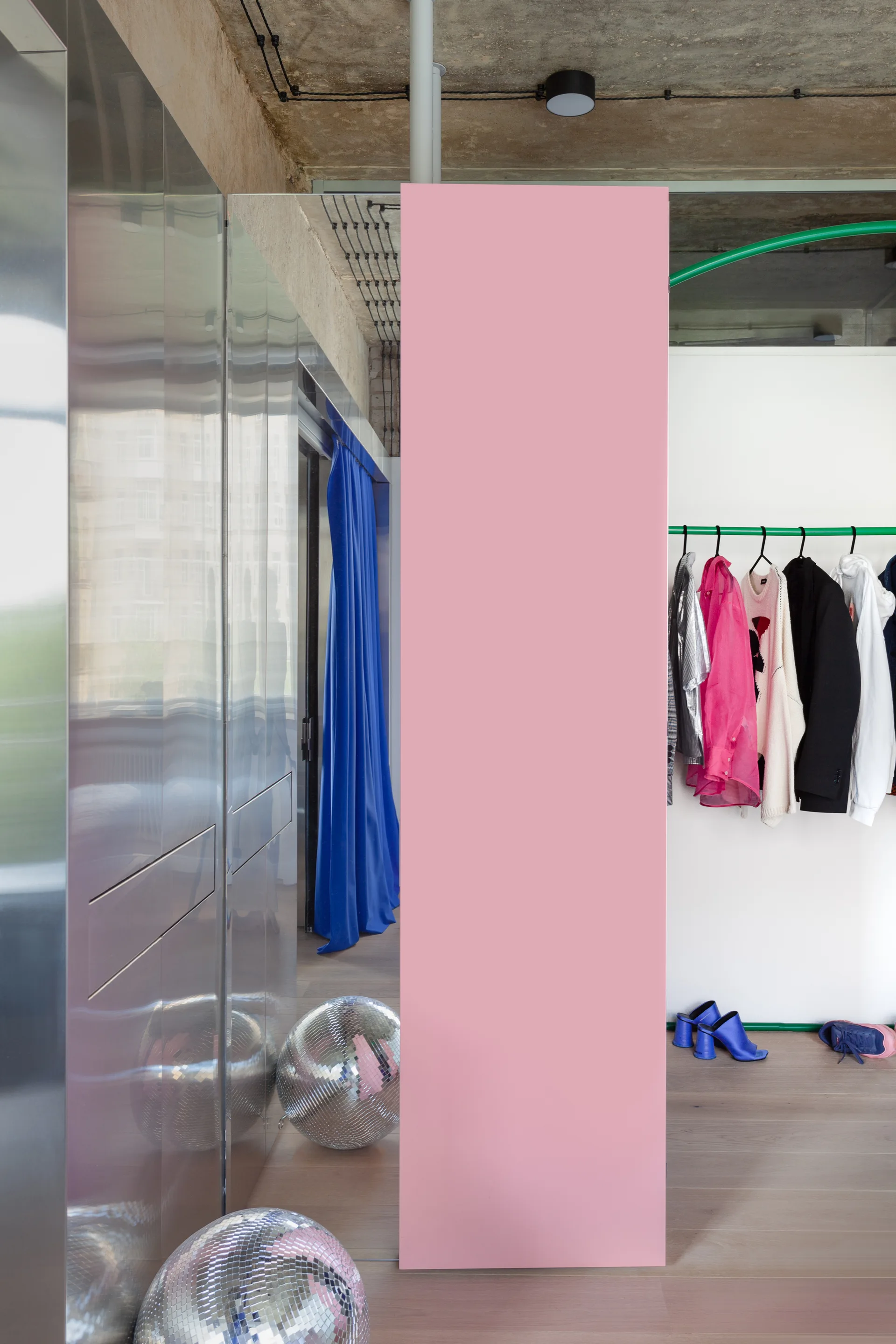 A modern dressing room features a bold pink partition, mirrored wardrobe cabinets, and a clothing rack beneath an exposed concrete ceiling.