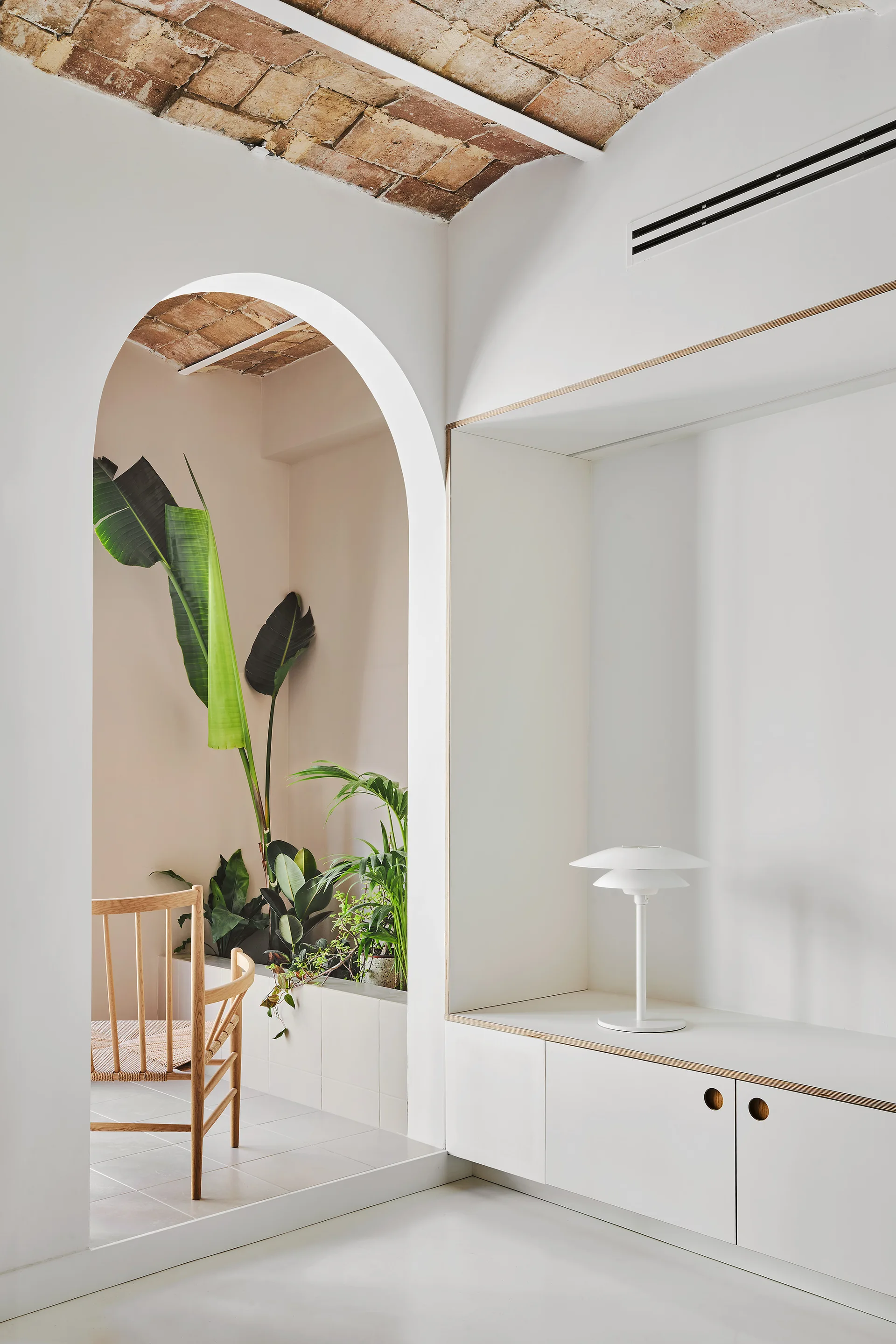 A minimalist white room features an arched passage leading to a plant-filled alcove beneath an exposed brick vaulted ceiling.