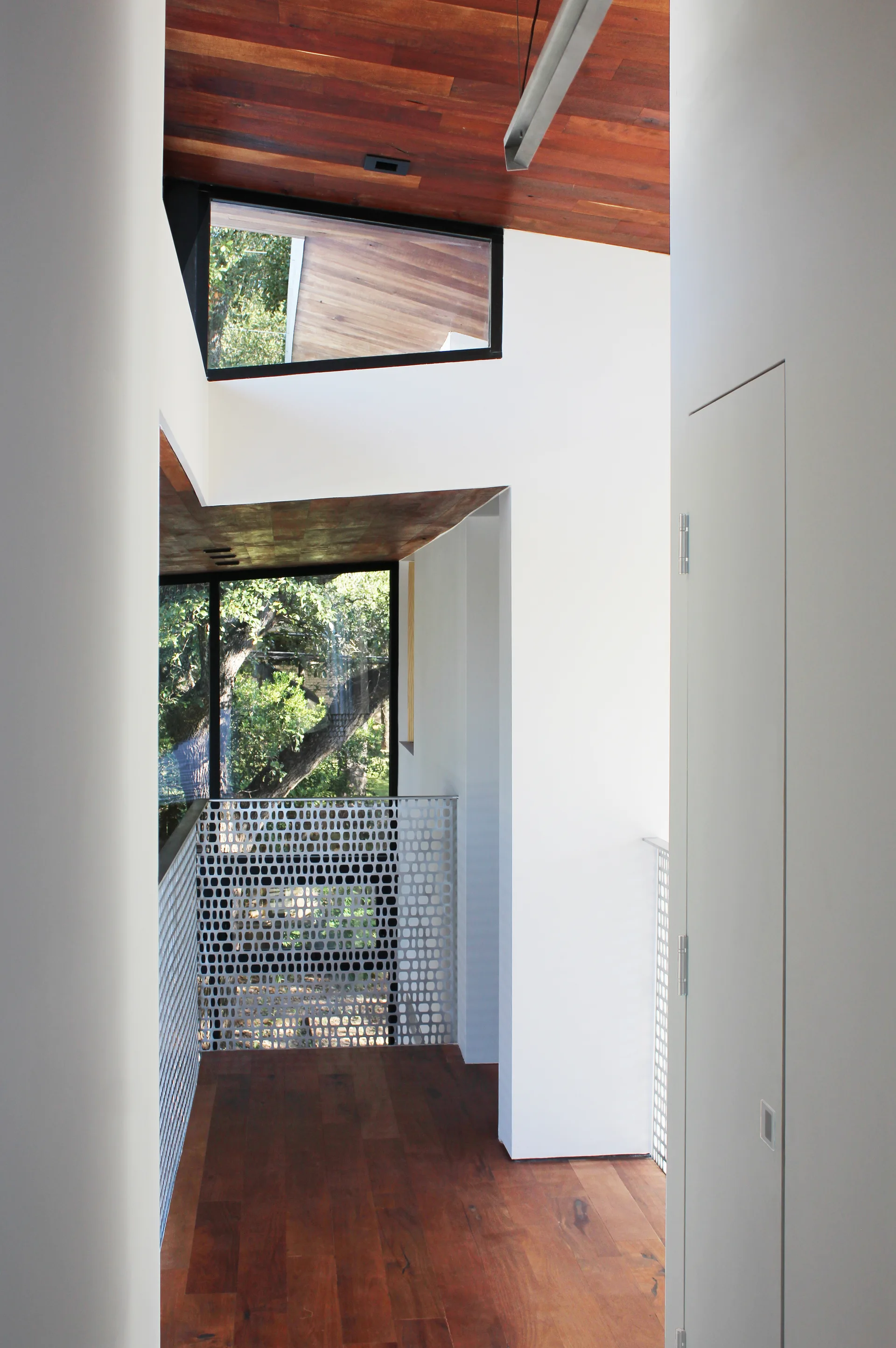 A modern hallway features rich wood flooring, a matching wood plank ceiling, clean white walls, and a patterned, perforated metal railing.
