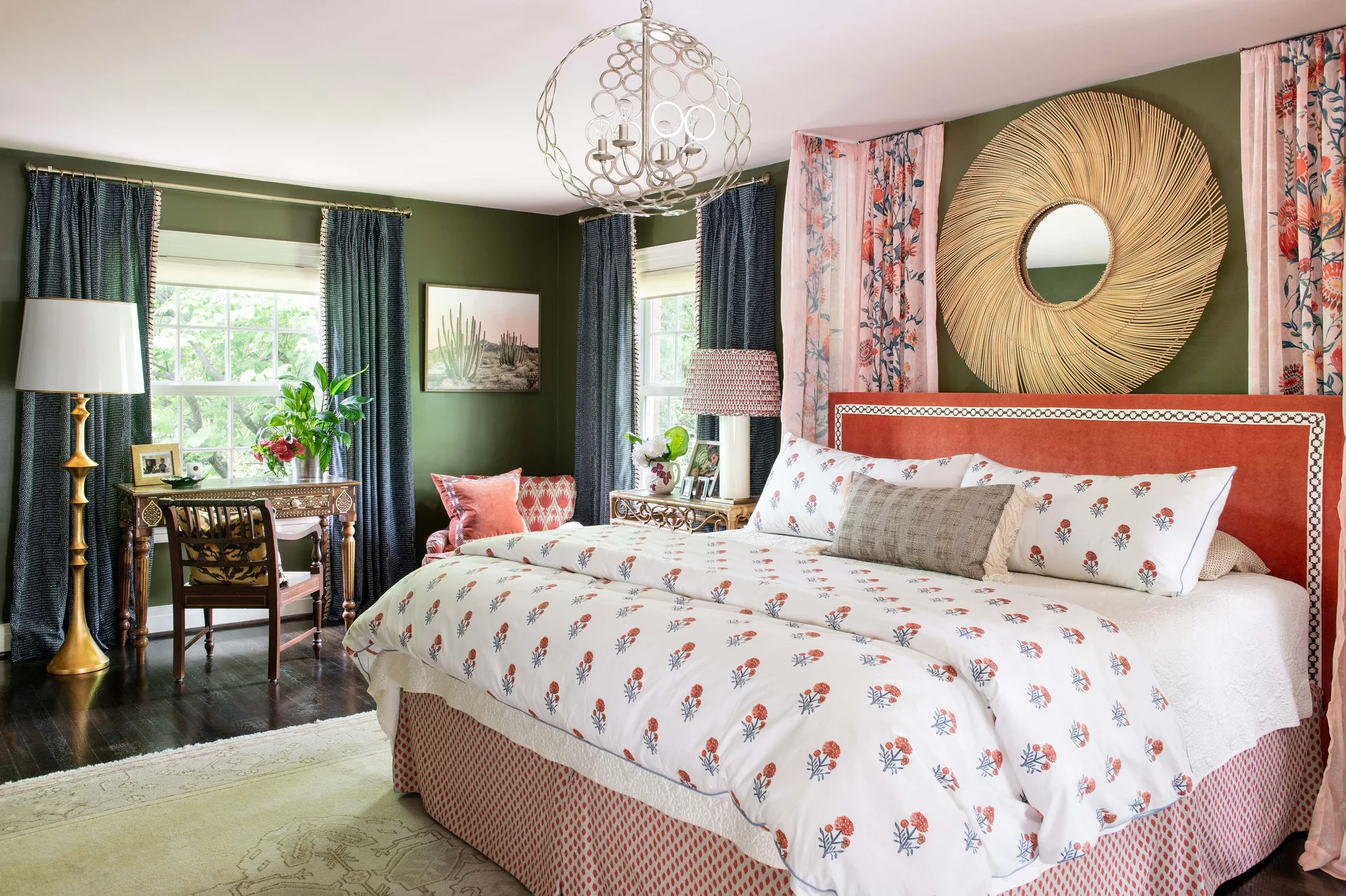An eclectic bedroom features olive green walls, an orange headboard with a rattan sunburst mirror, floral print bedding, and a wooden writing desk.