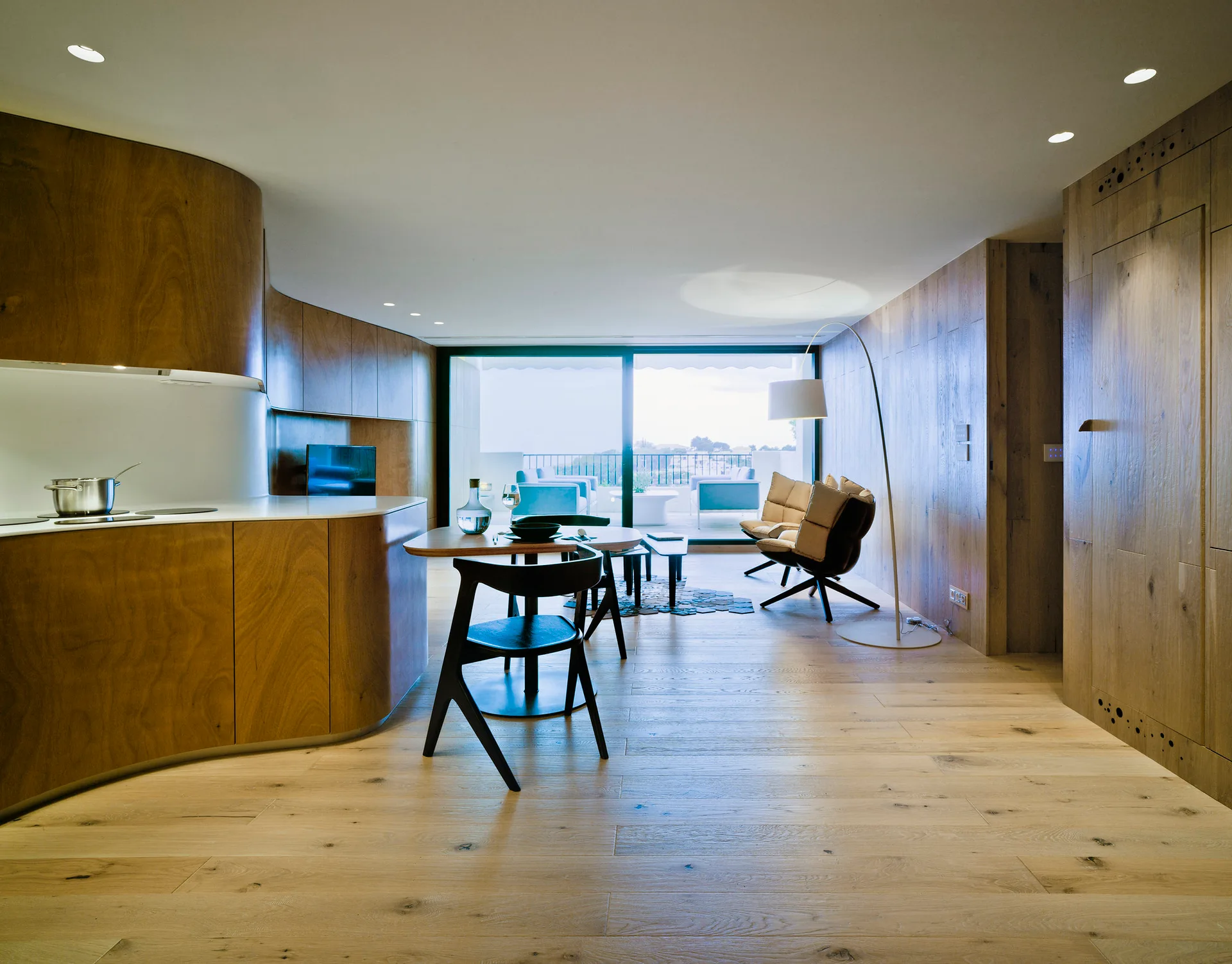 An open-plan living and kitchen area features curved wood cabinetry, a small dining table, wooden wall paneling, and sliding glass doors to a balcony.