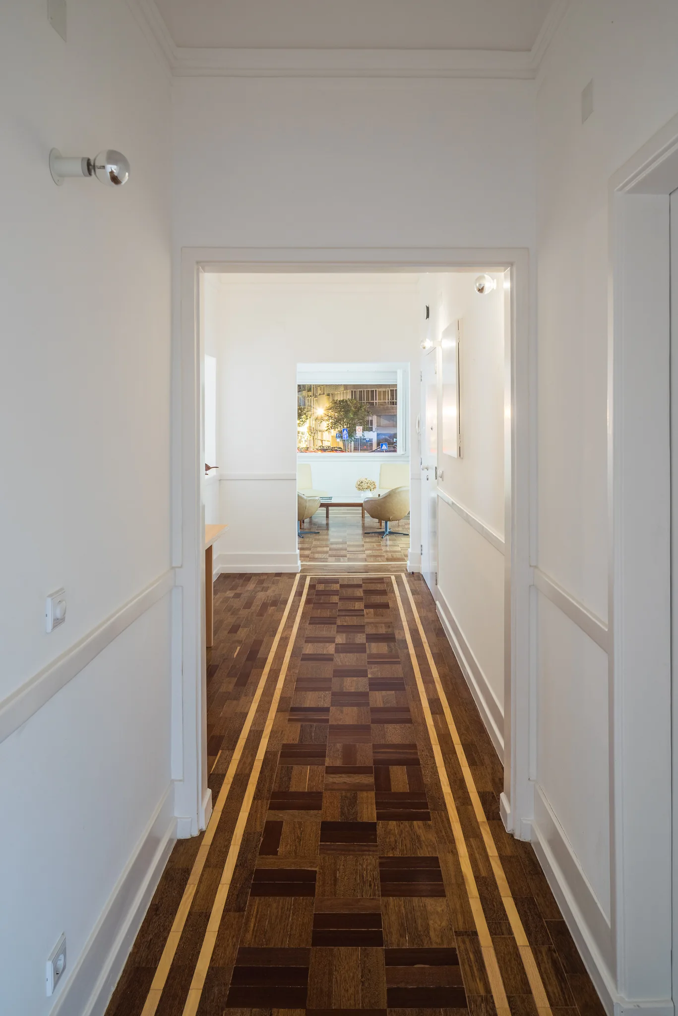 A narrow white hallway with wall molding and a geometric dark brown and tan parquet wood floor leading into a bright adjacent room.