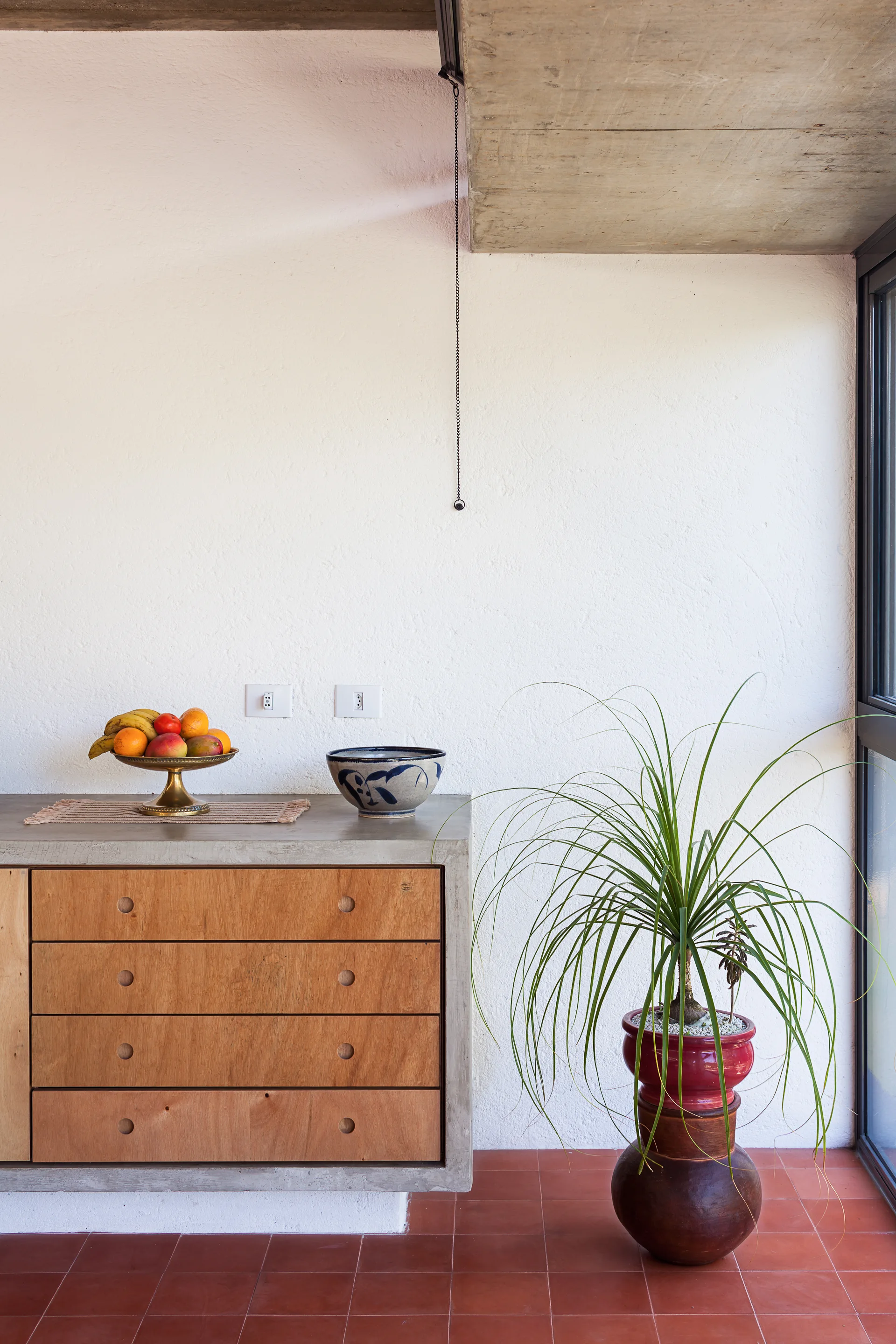 A minimalist built-in cabinet with plywood drawers and a concrete frame stands on a terracotta tile floor next to a potted houseplant.